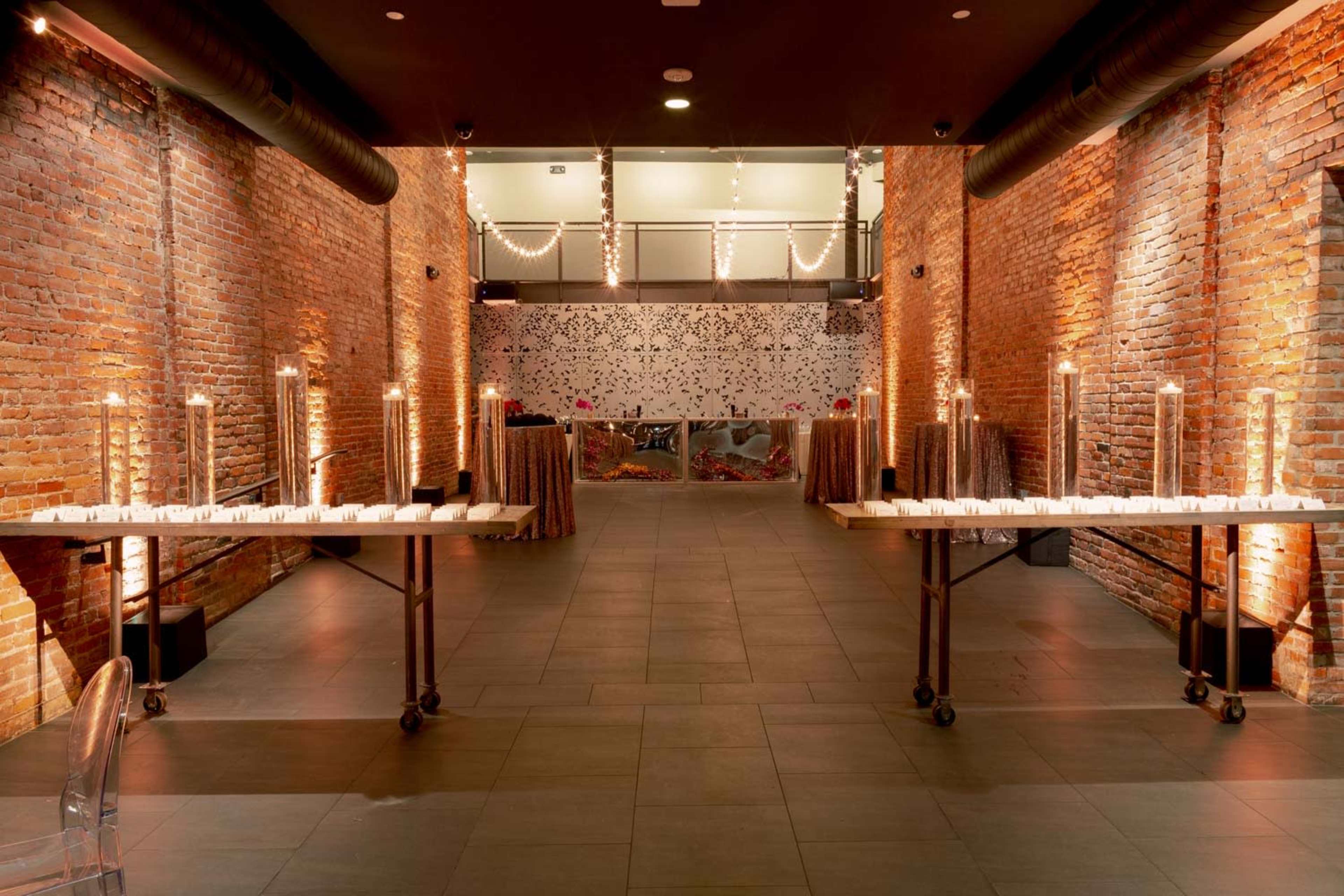 A spacious event venue with exposed brick walls, two tables arranged on either side adorned with candles, and decorative elements at the back.