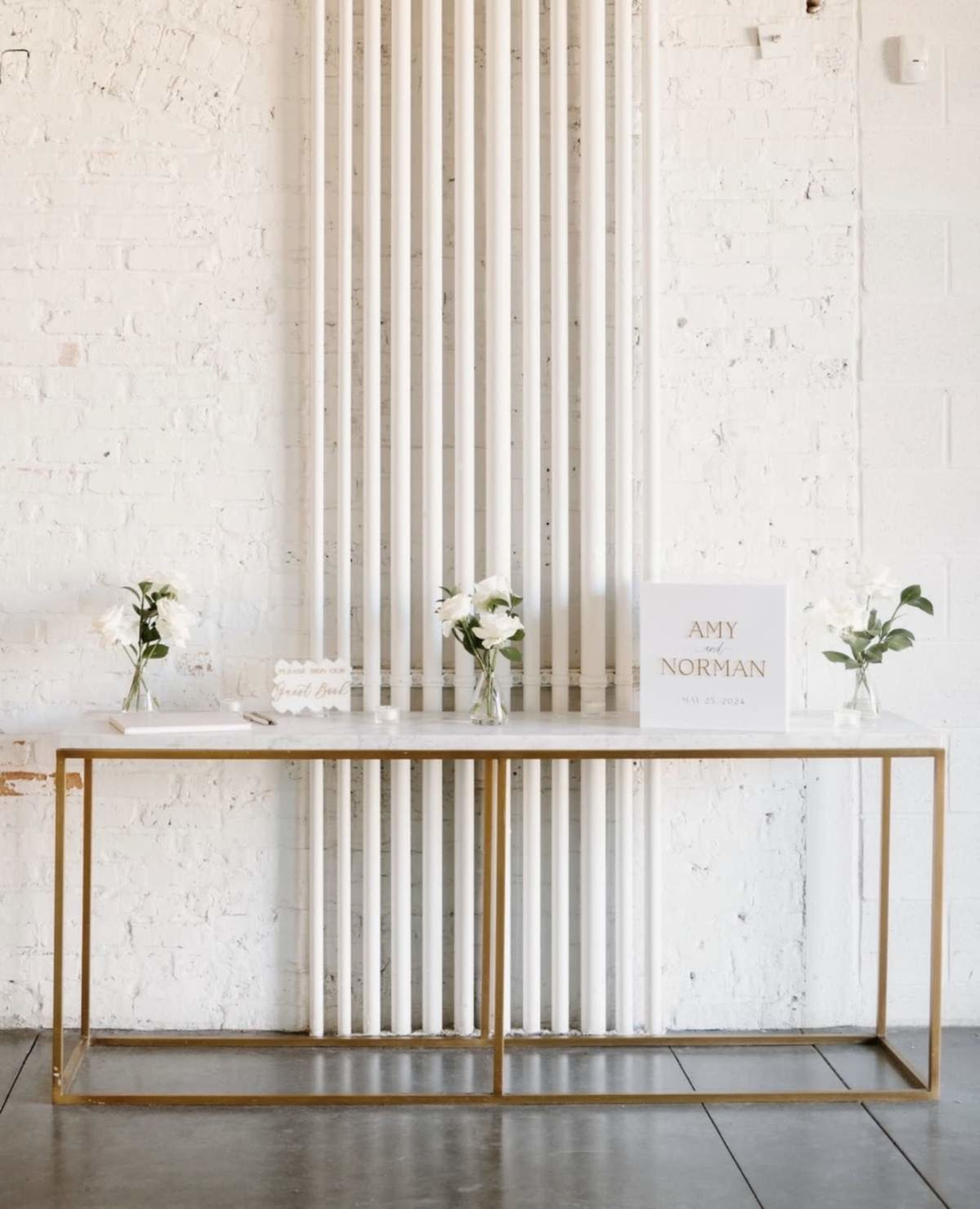 A long, gold-framed table against a white brick wall displays two vases with flowers and a sign reading "AMY NORMAN."