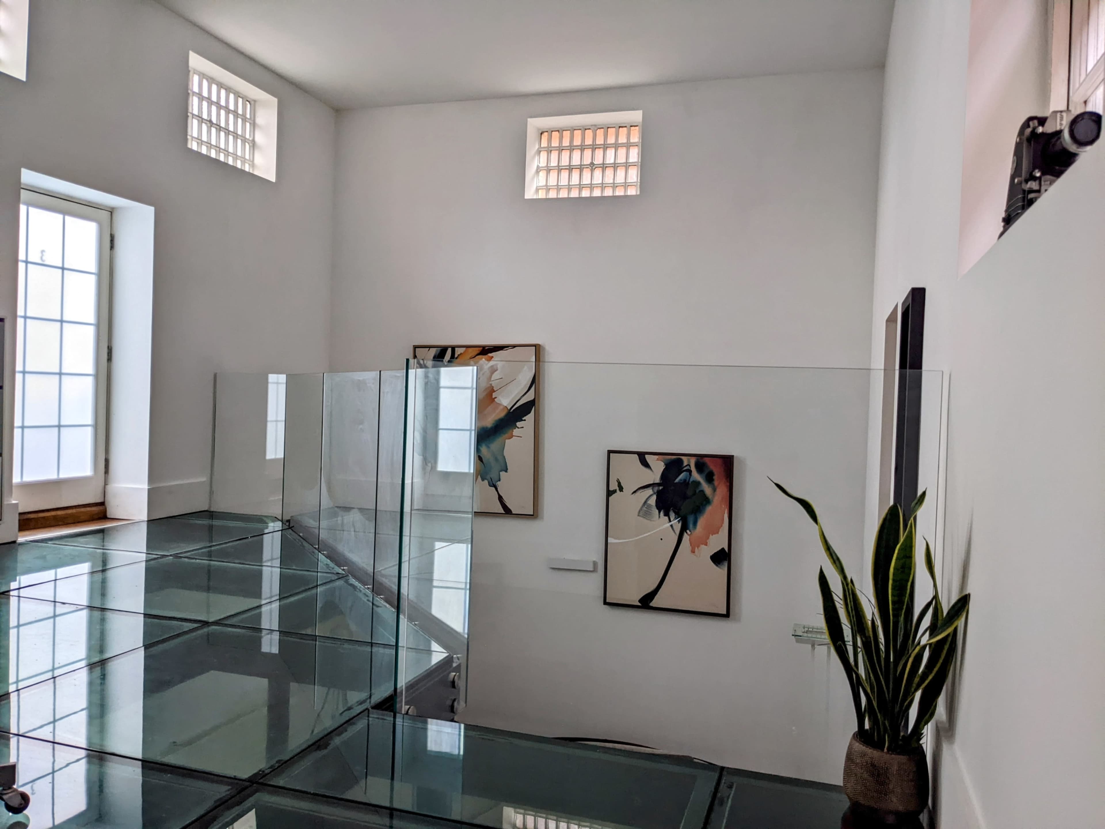 A modern interior space with glass walls, art pieces on the white walls, and a potted plant in the corner.