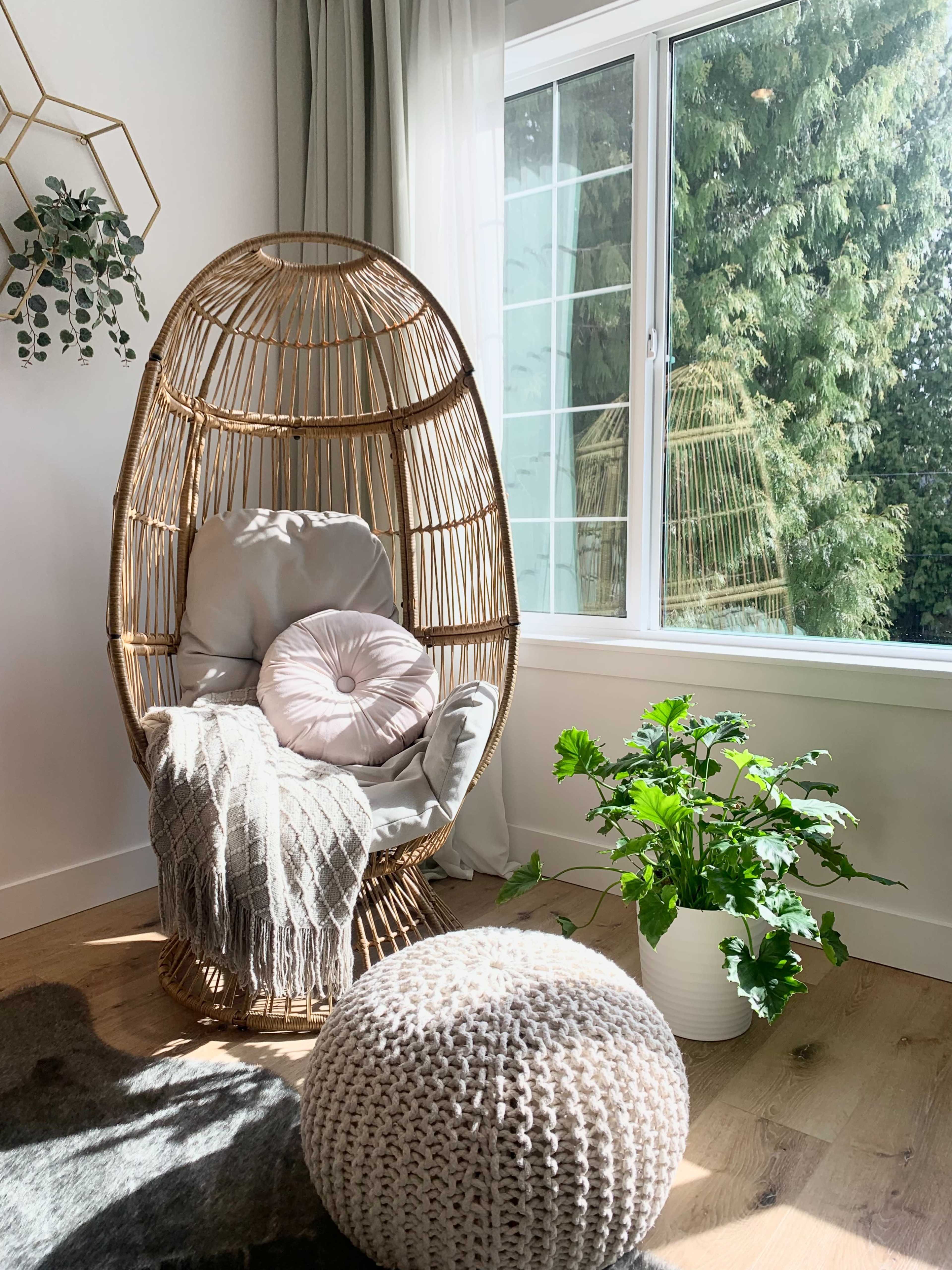 A cozy corner featuring a wicker hanging chair, a decorative cushion, a knit pouf, and a potted plant by a large window with greenery outside.