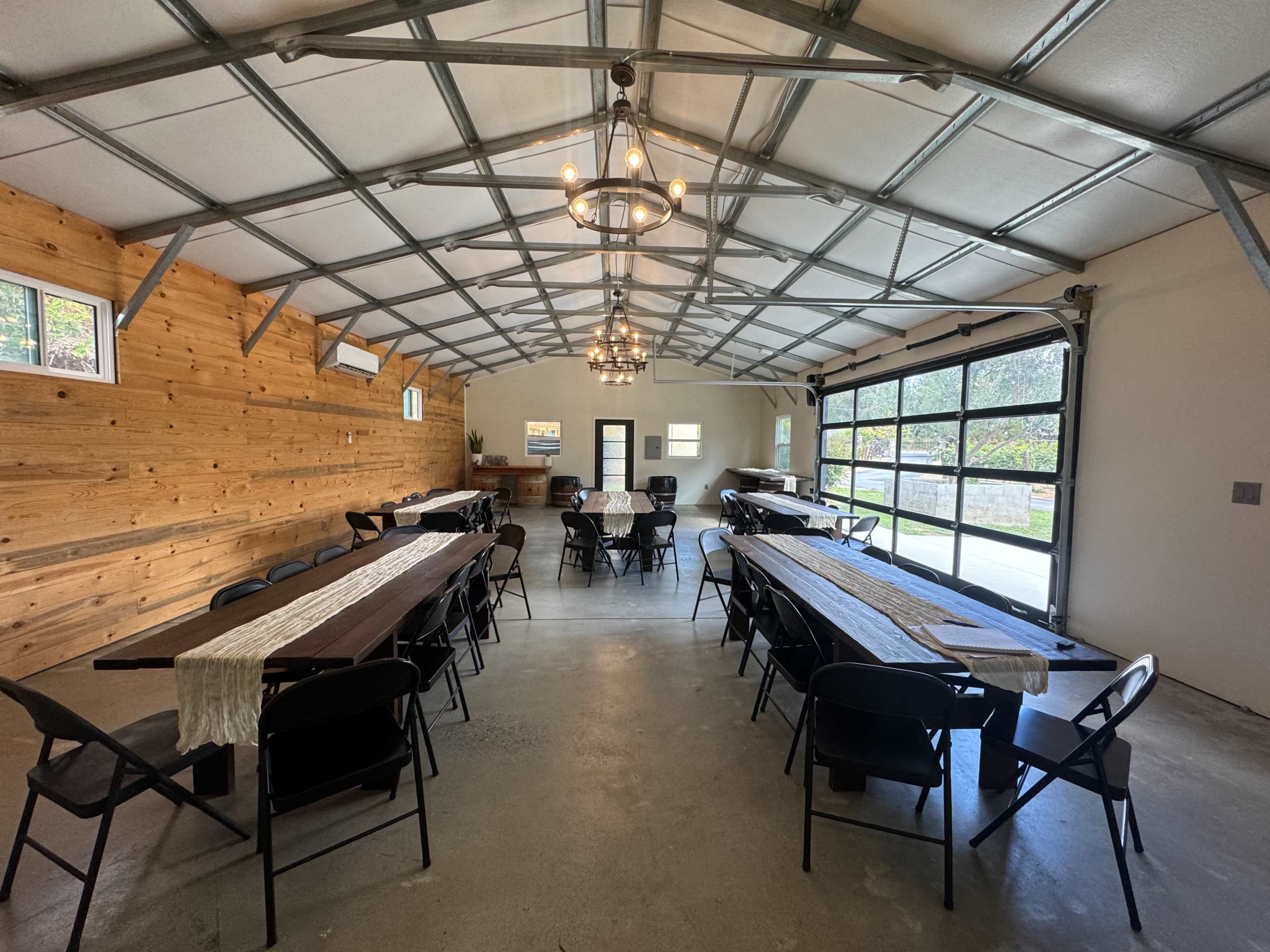 The image shows a spacious, well-lit event room with several long tables arranged in rows, wooden walls, and large windows, complemented by industrial-style lighting fixtures.
