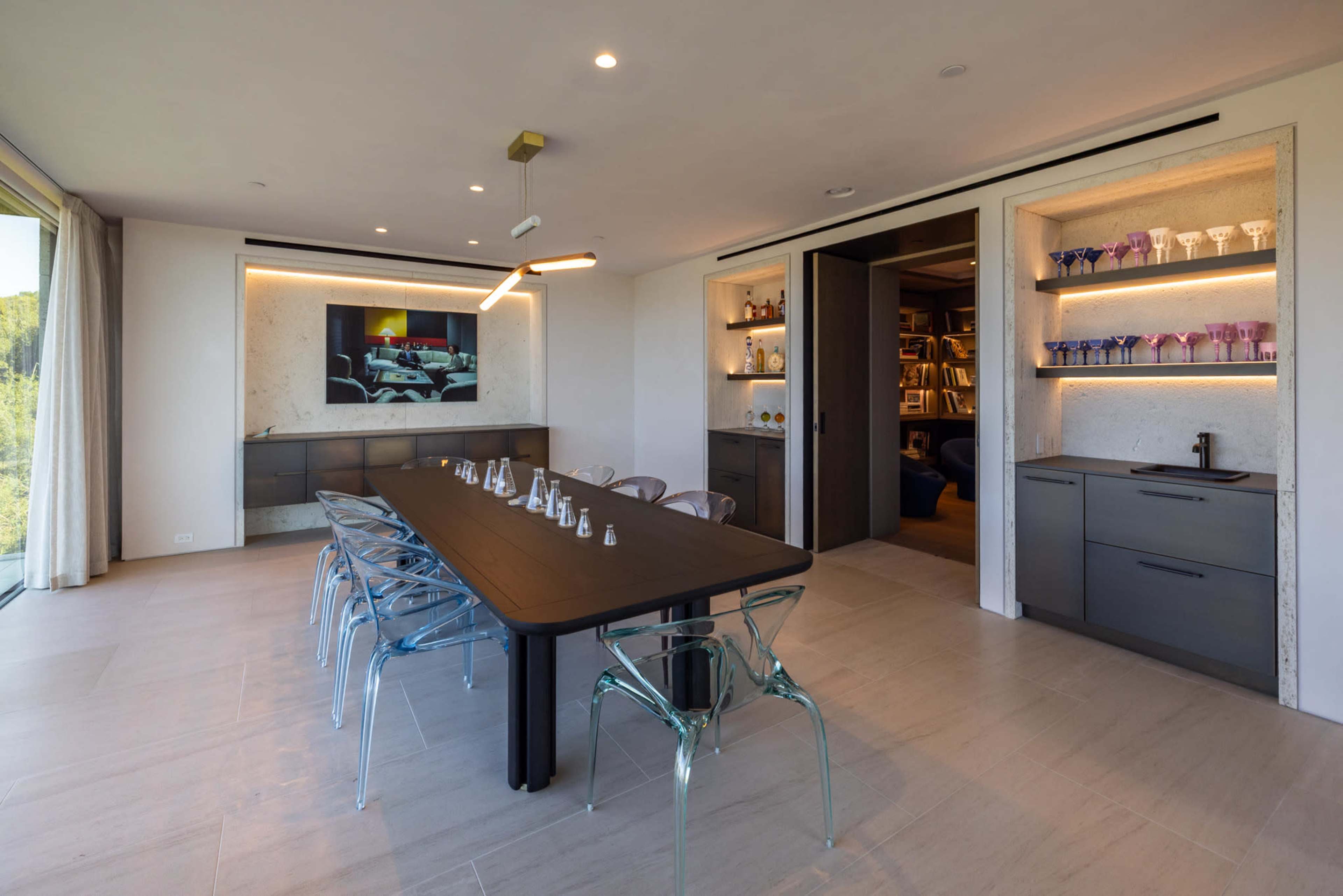 The image shows a modern dining room with a rectangular black table surrounded by transparent chairs, a wall-mounted television, and shelves displaying glassware.