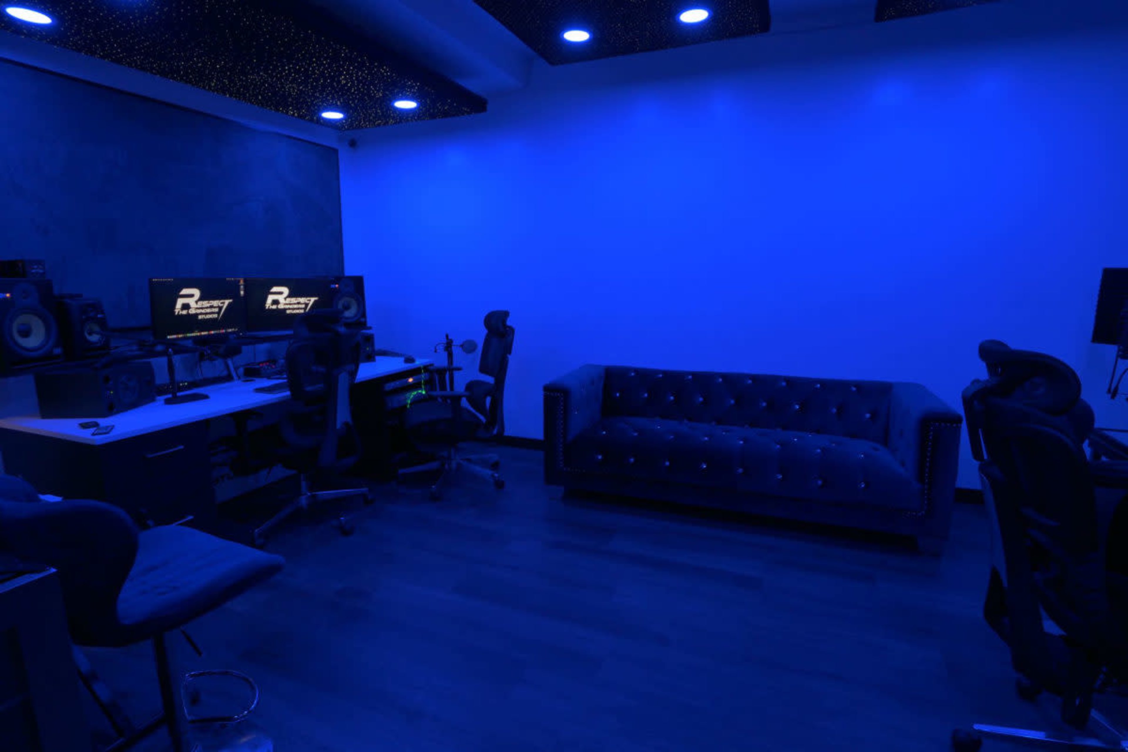 The image shows a dimly lit music studio featuring a blue-toned interior with a desk, computer monitors, and a sofa.