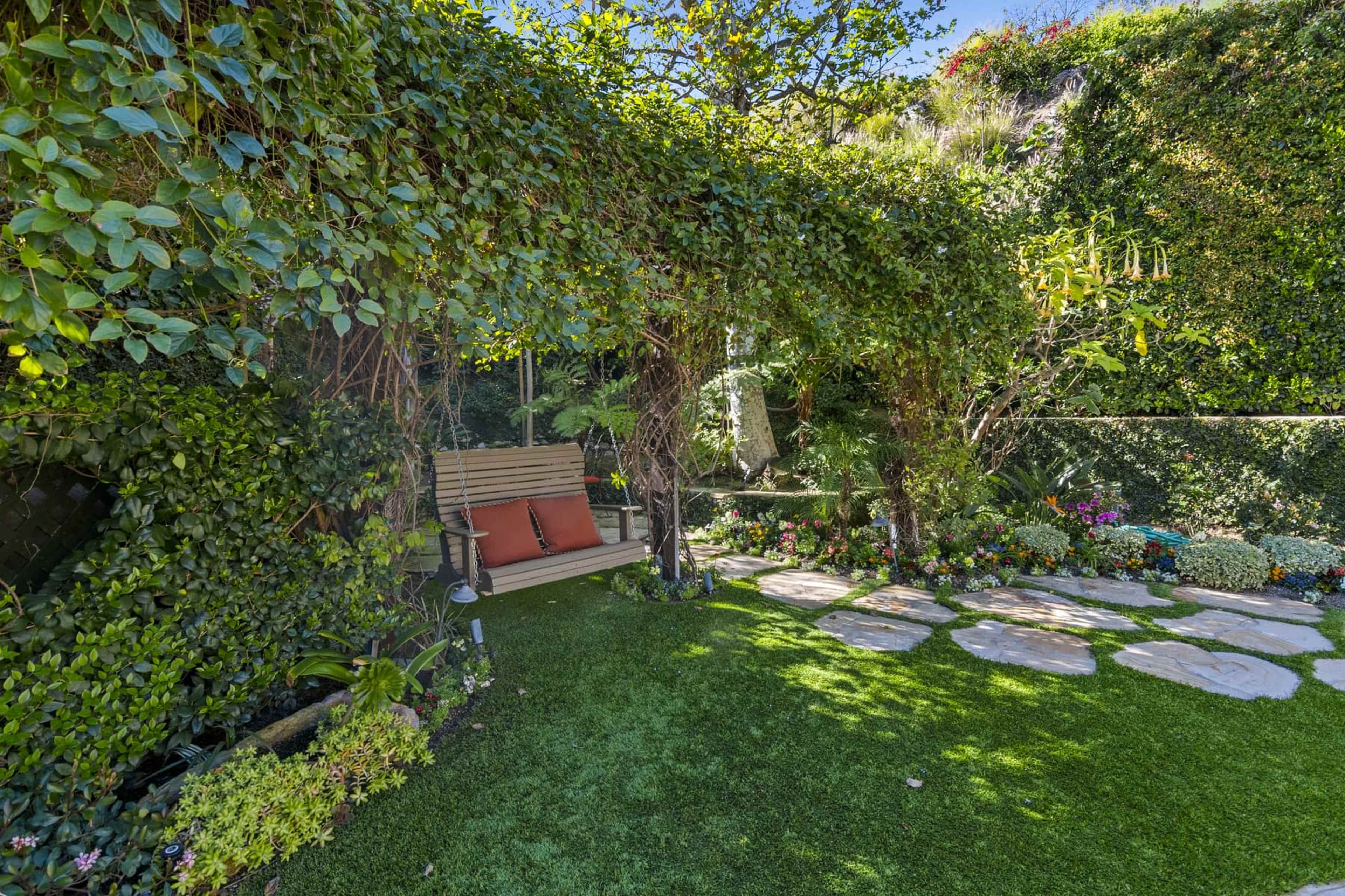 A wooden swing with two red cushions hangs beneath a lush green canopy in a well-tended garden filled with flowers and stone pathways.