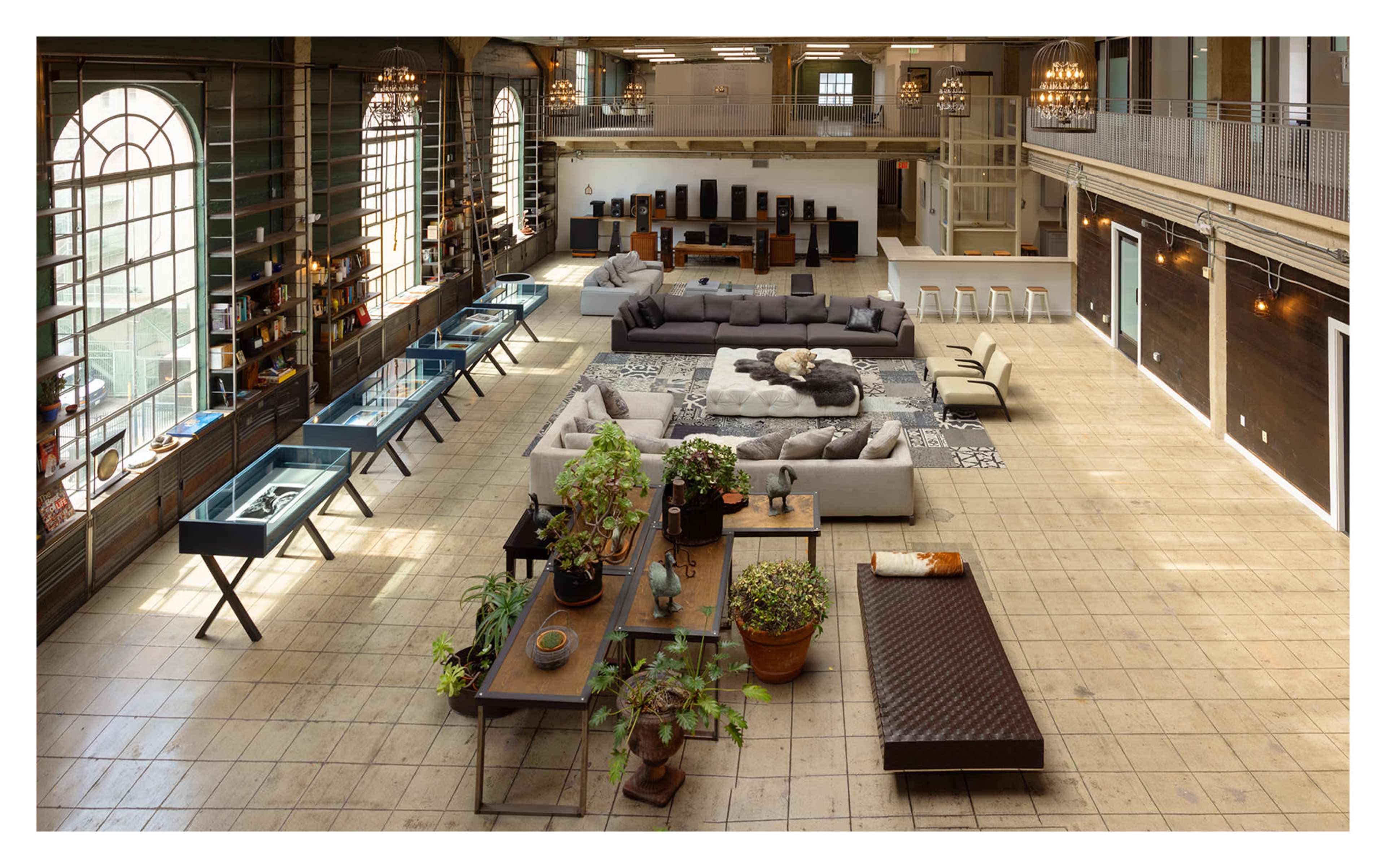 The image shows a spacious, modern interior with large windows, featuring a mixed arrangement of seating areas, tables, plants, and decorative displays.