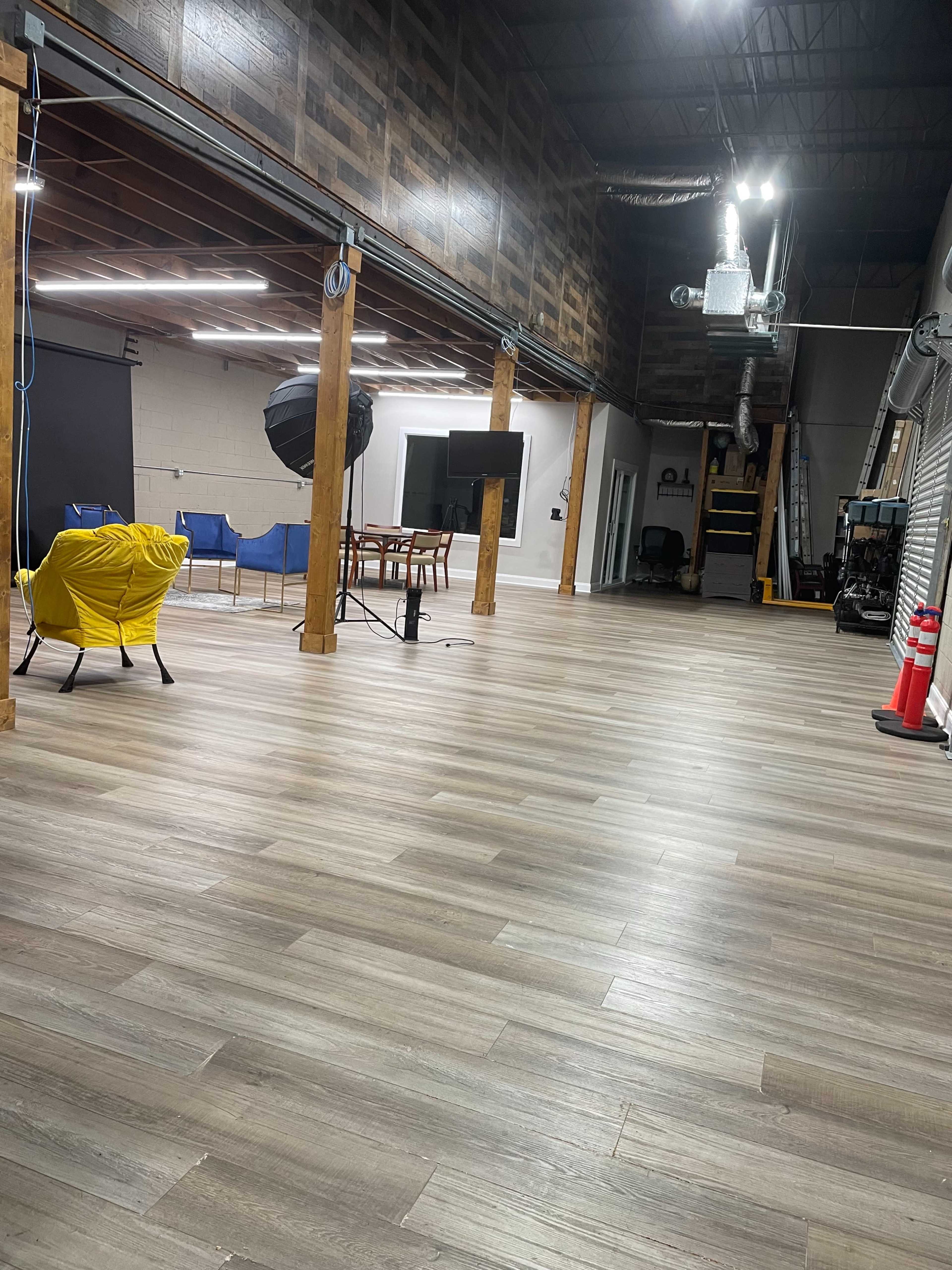 A spacious interior with wooden beams, modern flooring, and various furniture items including chairs, a table, and lighting equipment.