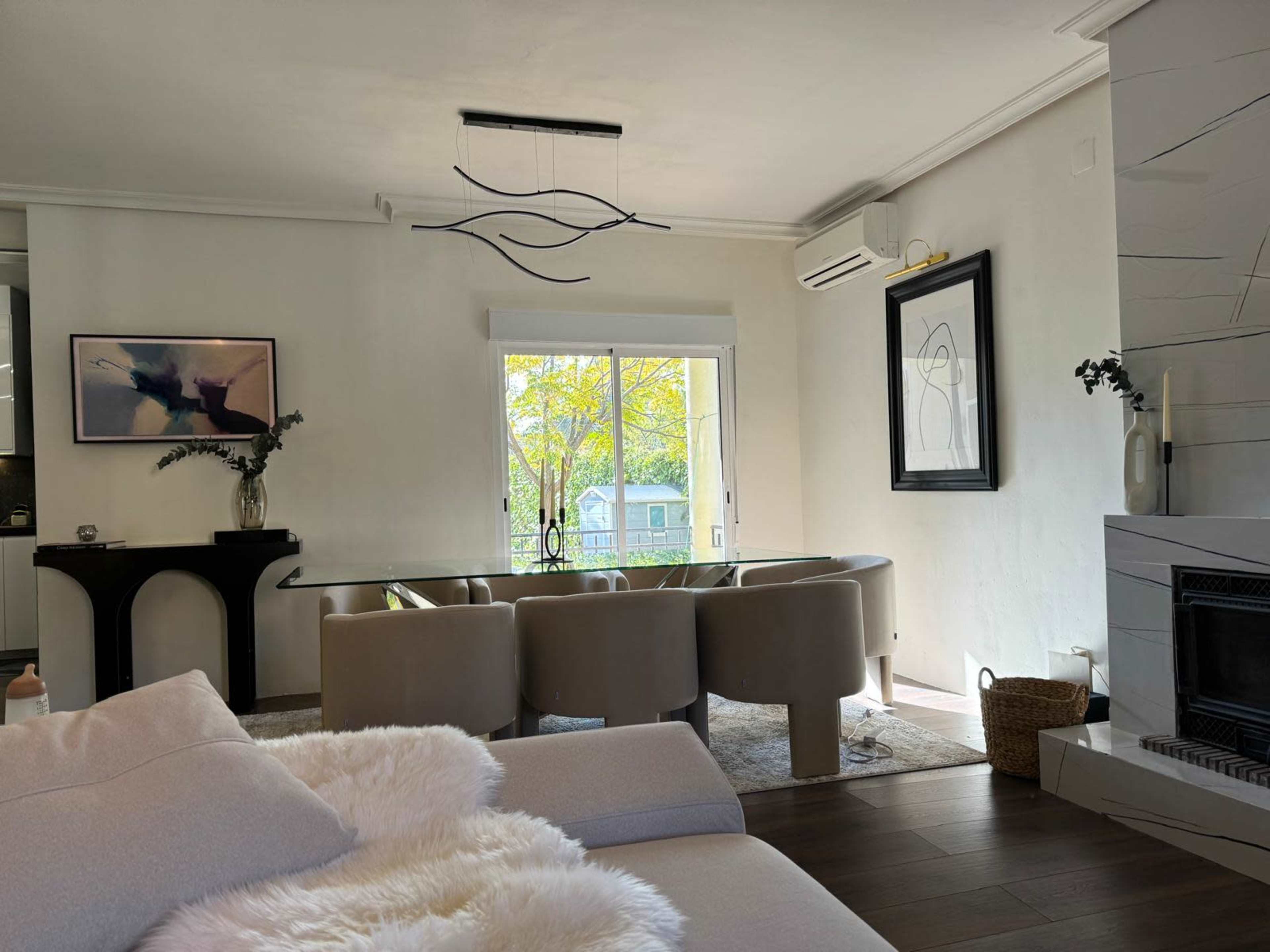 The image shows a modern living room with a light-colored sofa, a glass dining table surrounded by upholstered chairs, and large windows letting in natural light.
