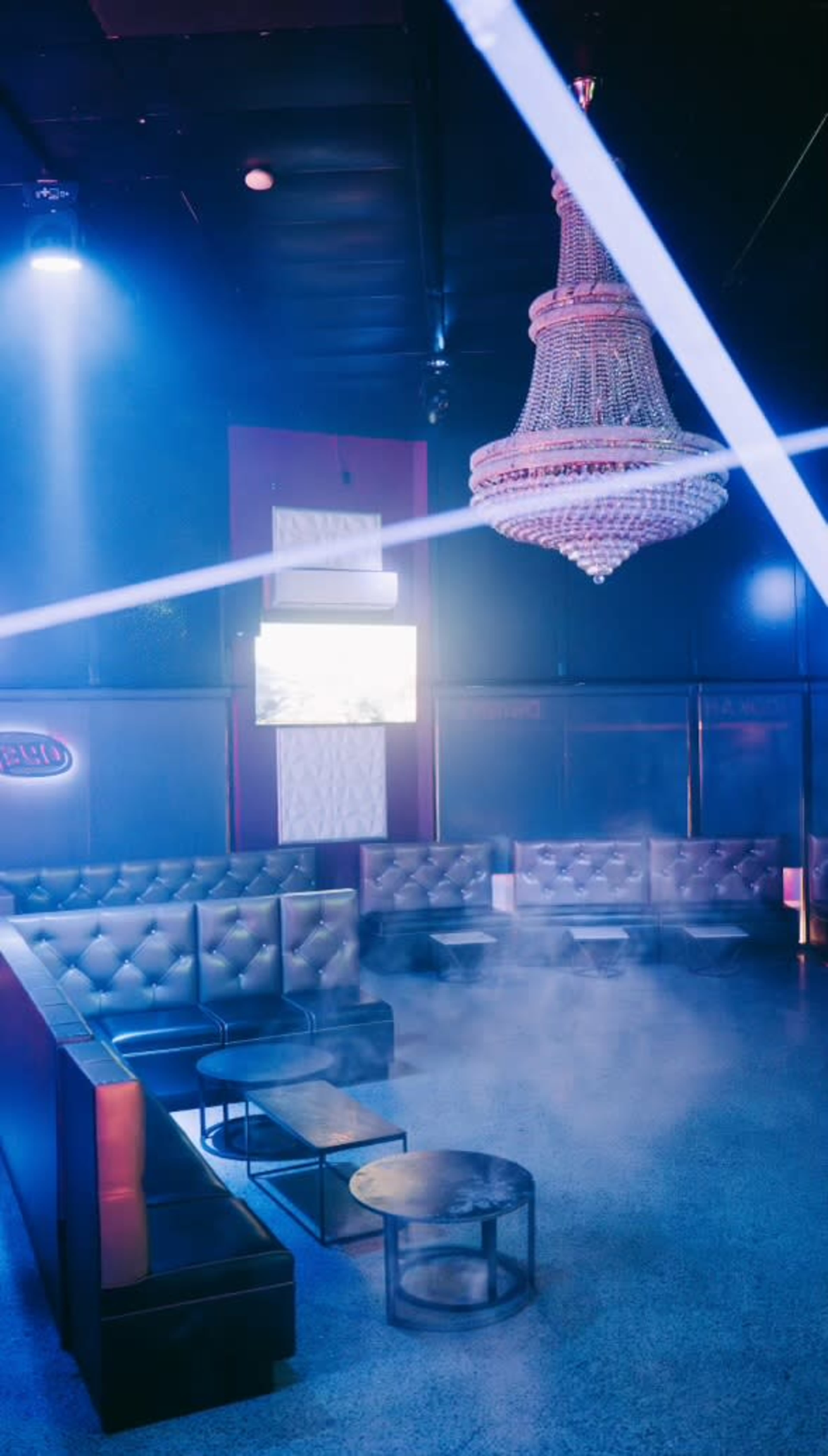A dimly lit lounge features leather seating, a chandelier, and a misty atmosphere enhanced by blue lighting.