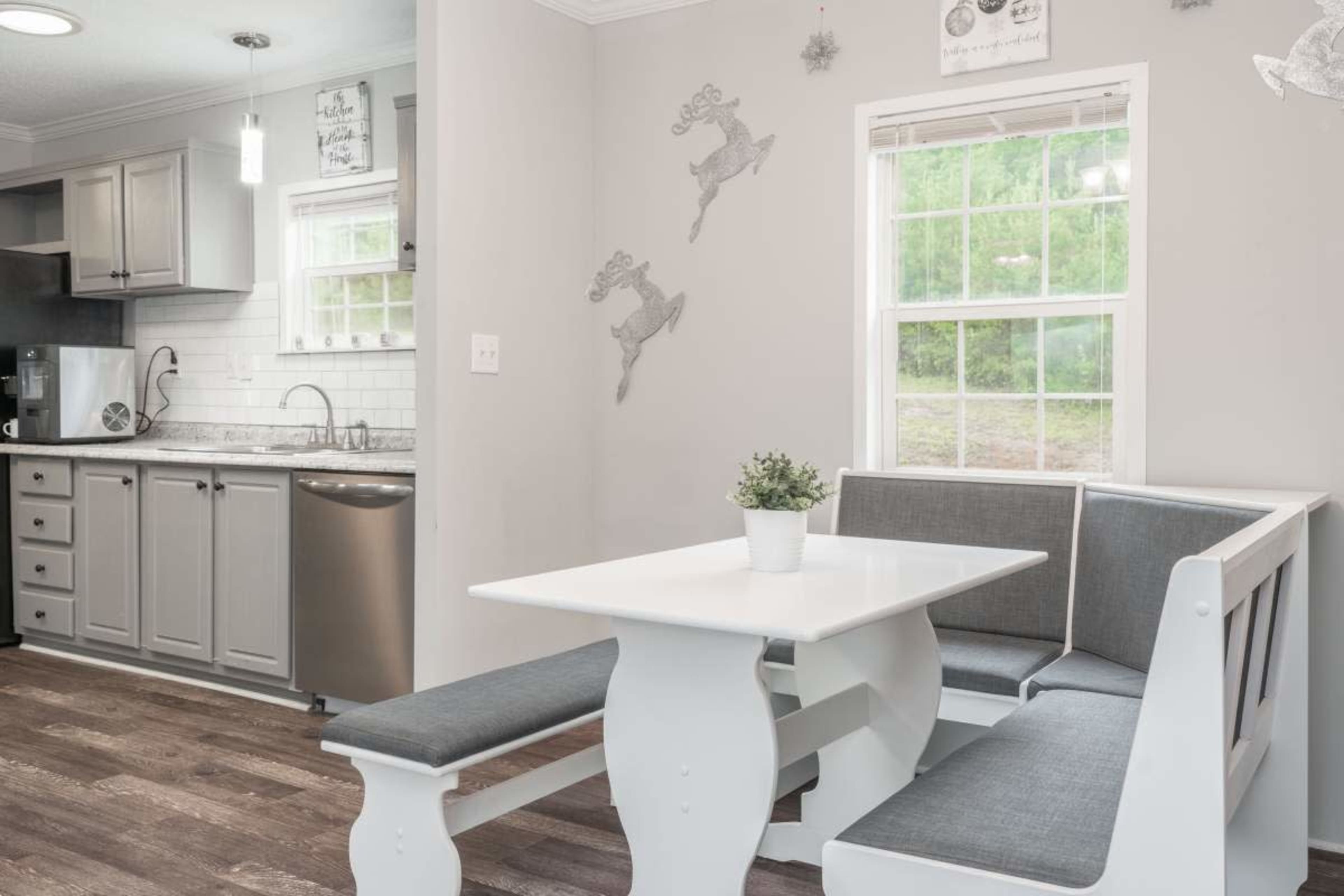 The image shows a modern kitchen with gray cabinetry, white walls, and a dining area featuring a white table and gray upholstered benches next to a window.