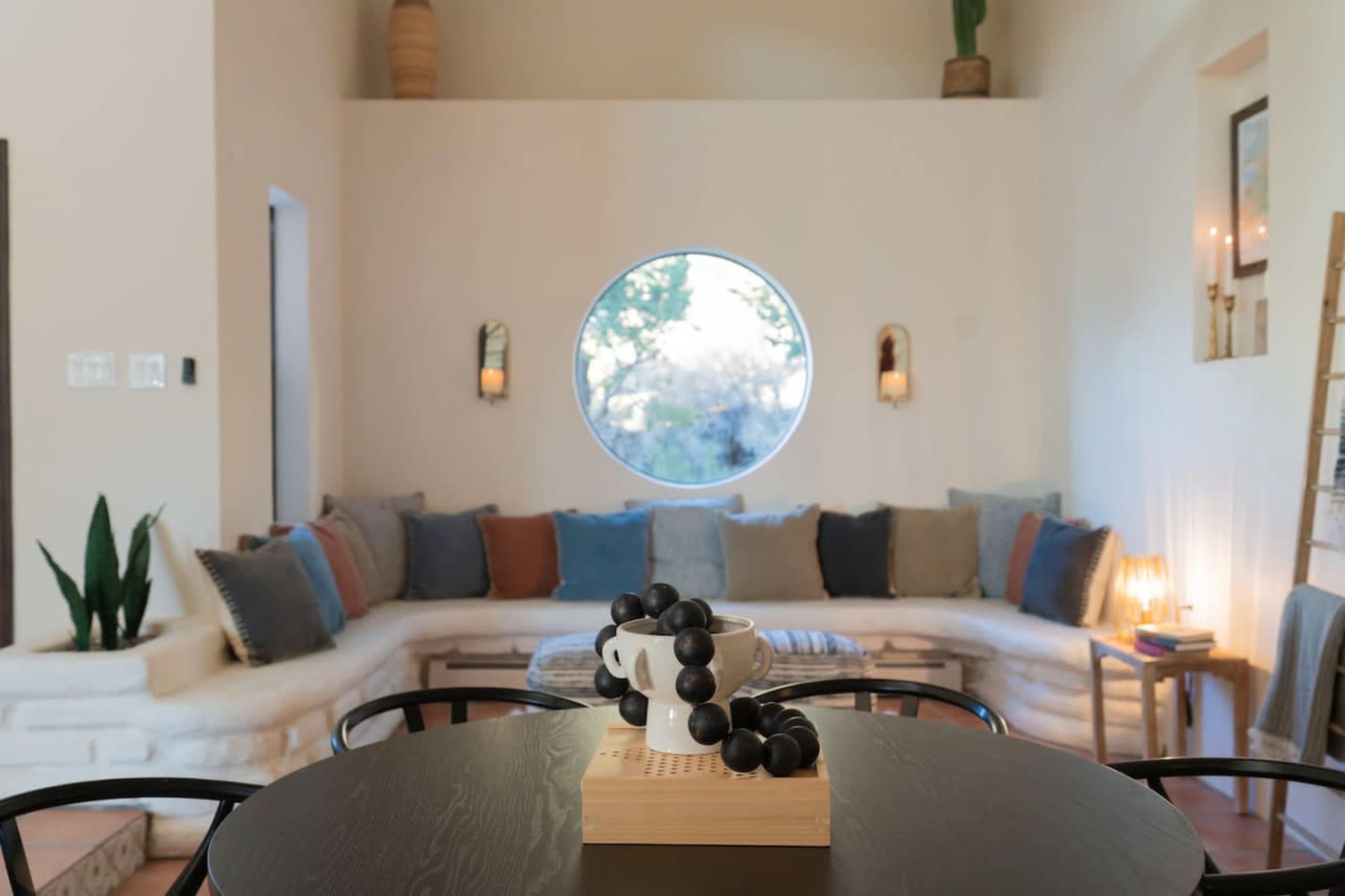 A cozy living area features a circular window, plush seating adorned with various cushions, and a round dining table with a decorative centerpiece.