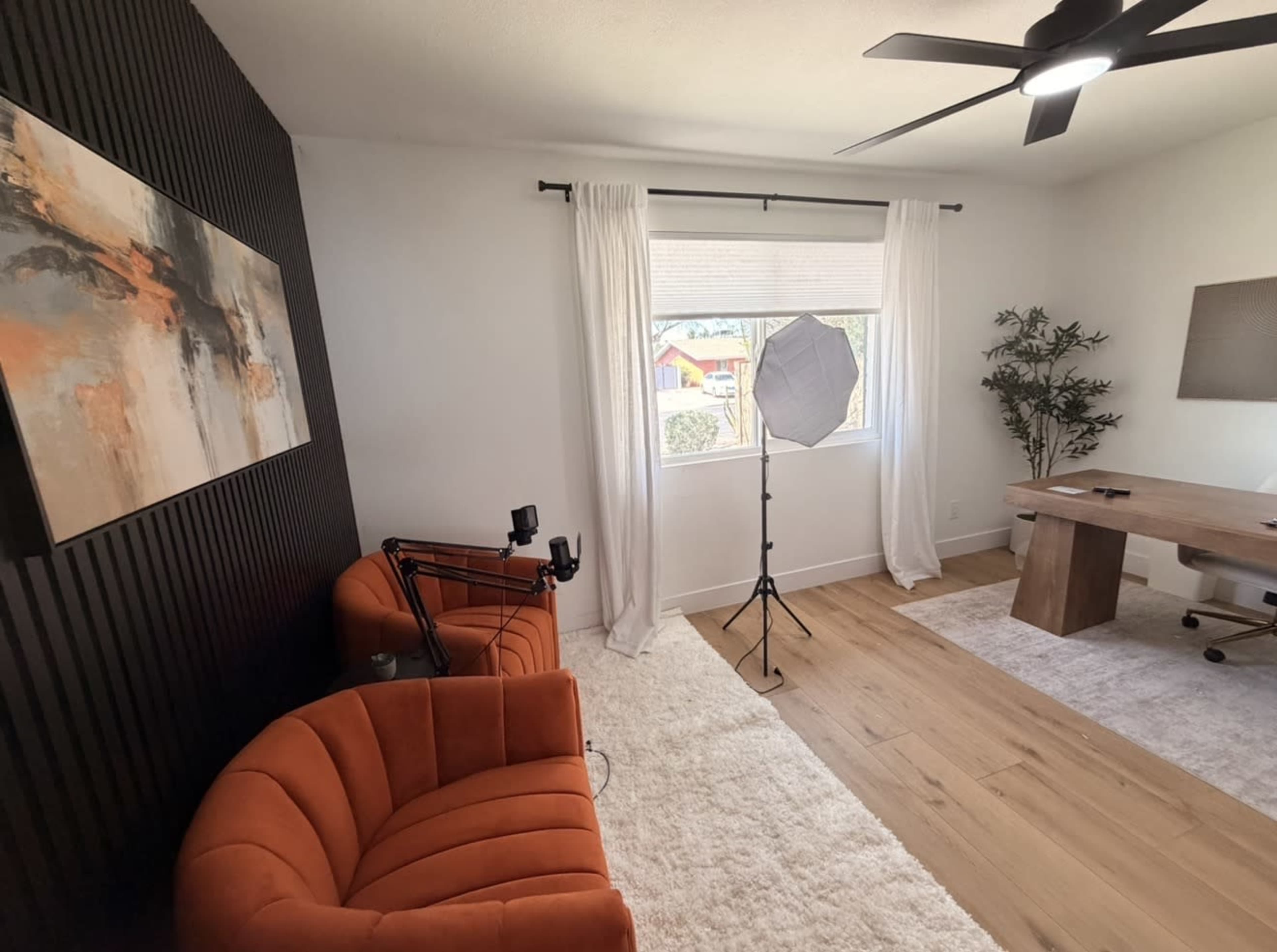 The room features an orange upholstered sofa, a wooden desk with a chair, and a large window with sheer curtains illuminating the space.
