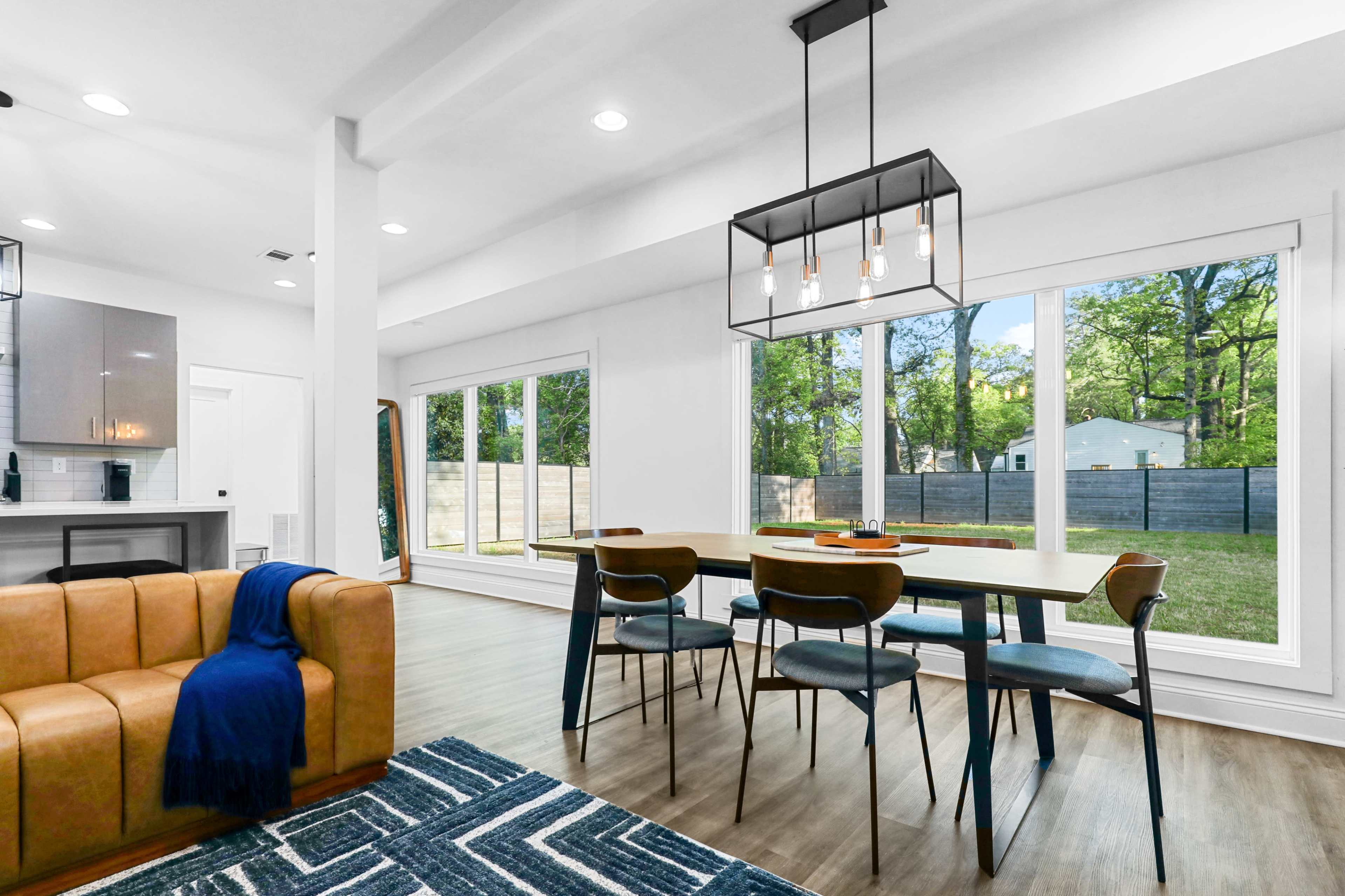 A modern dining area features a large table surrounded by chairs, with a brown sofa and large windows overlooking a green backyard.