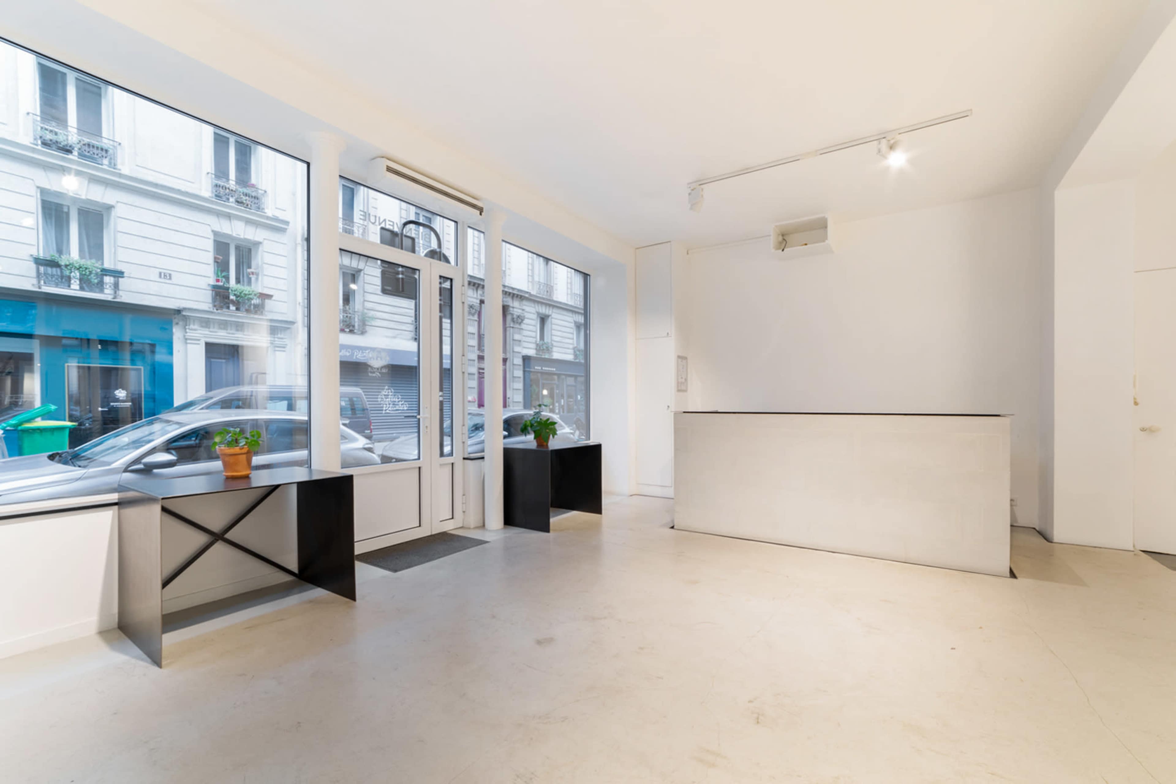 The image shows a minimalist interior of a retail space with large windows, a low reception desk, and two black display tables.