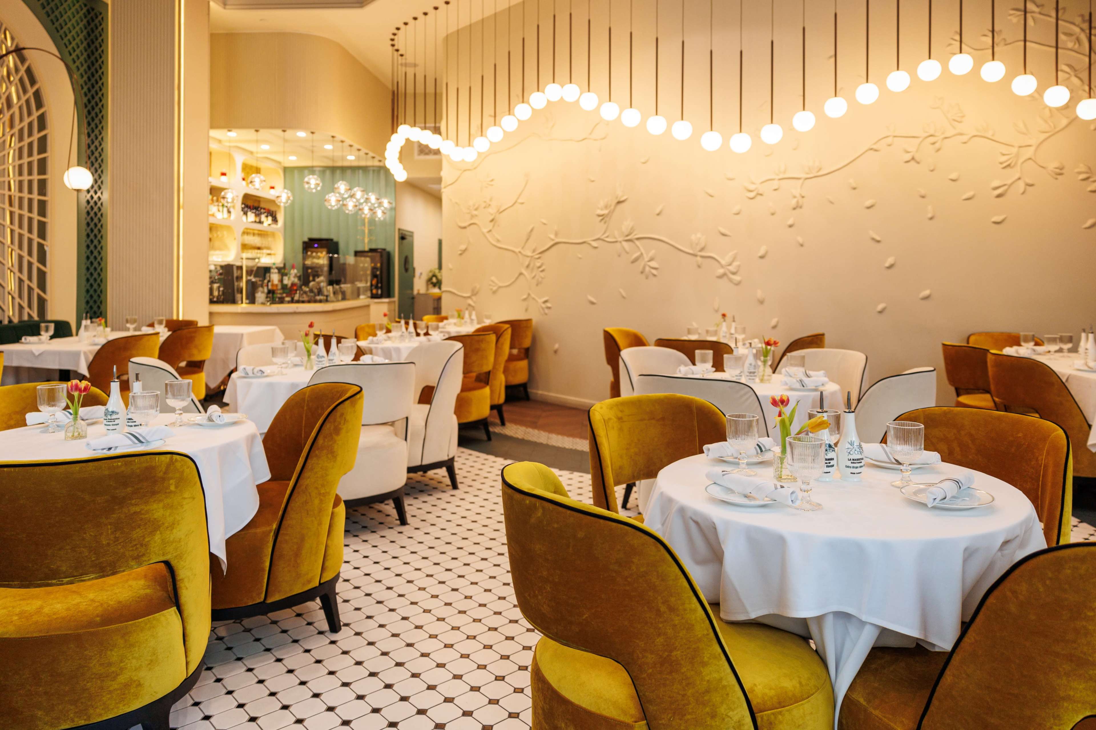 The image shows a stylish restaurant interior with round tables set for dining, surrounded by yellow upholstered chairs and features decorative wall elements and soft lighting.