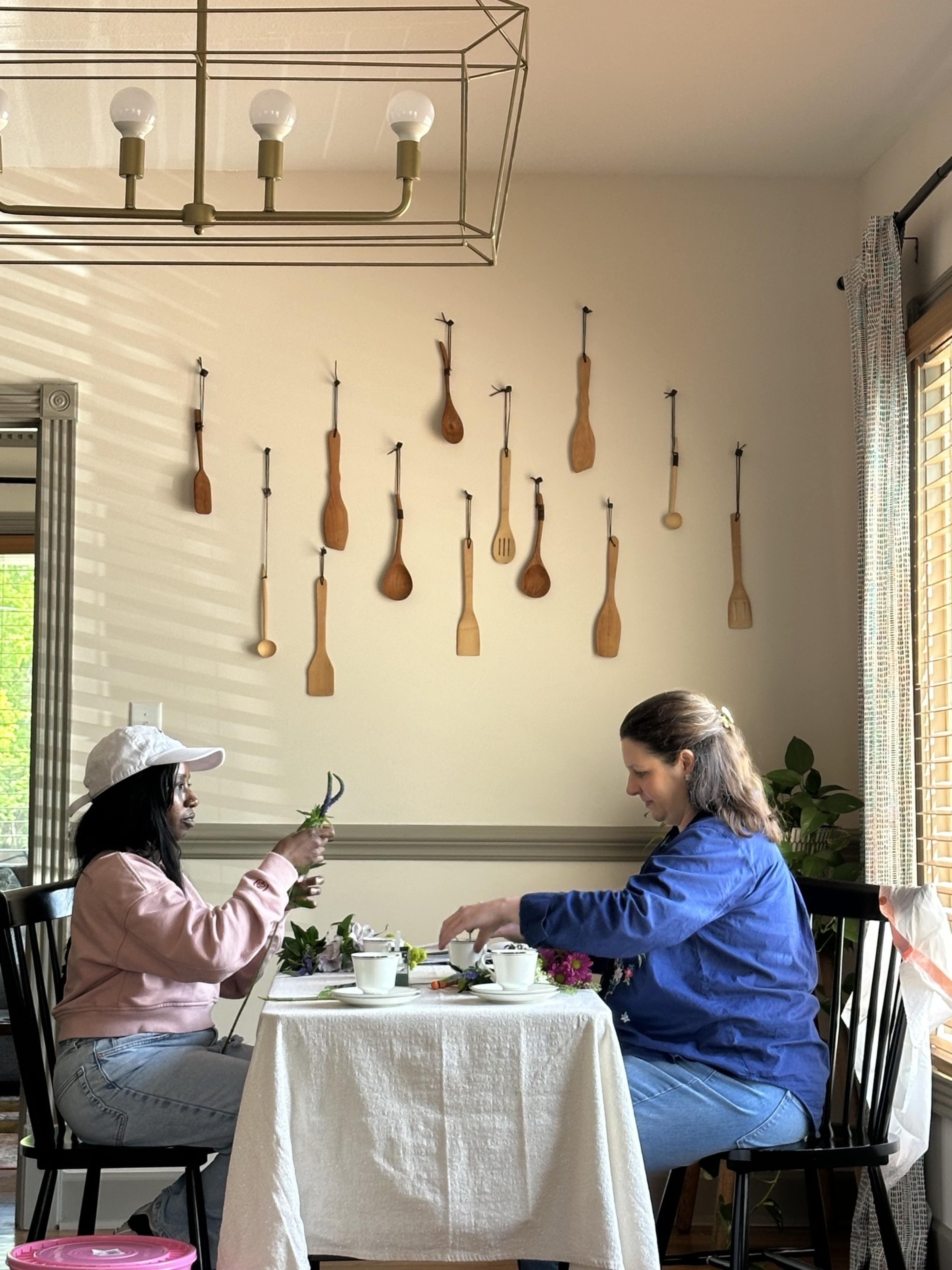 Two women sit at a table in a bright room decorated with wooden utensils on the wall, engaged in a floral arrangement activity.