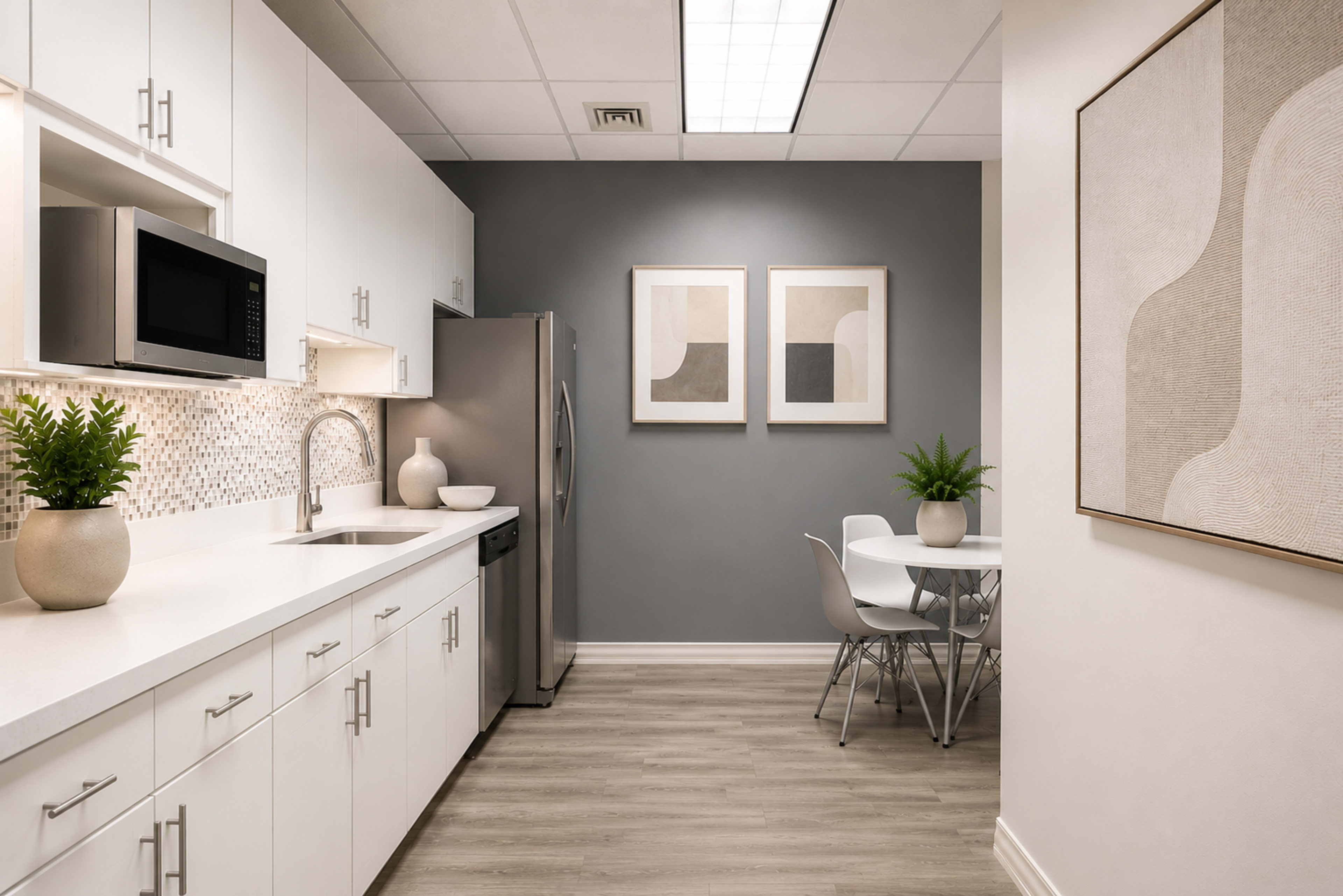 A modern kitchen features white cabinetry, stainless steel appliances, and a dining area with a round table and chairs, against a backdrop of gray walls and framed artwork.
