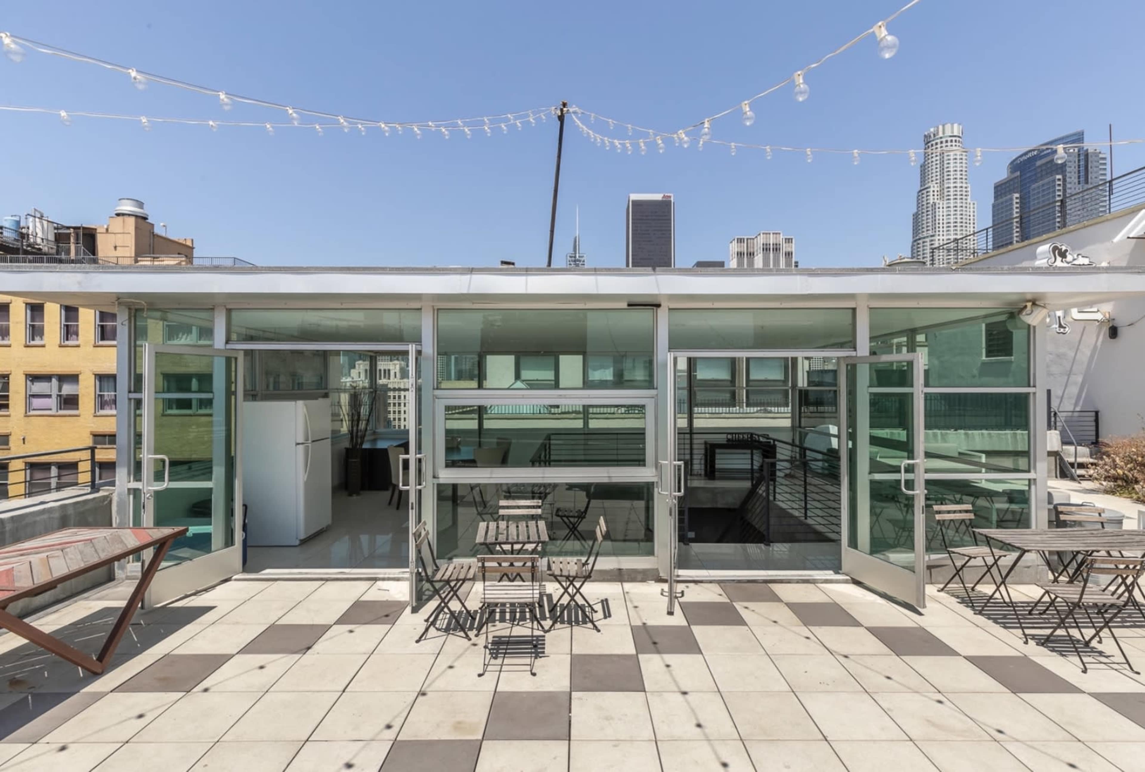A rooftop space with glass walls, stone tiles, and metal chairs and tables, set against a backdrop of city skyscrapers under a clear blue sky.
