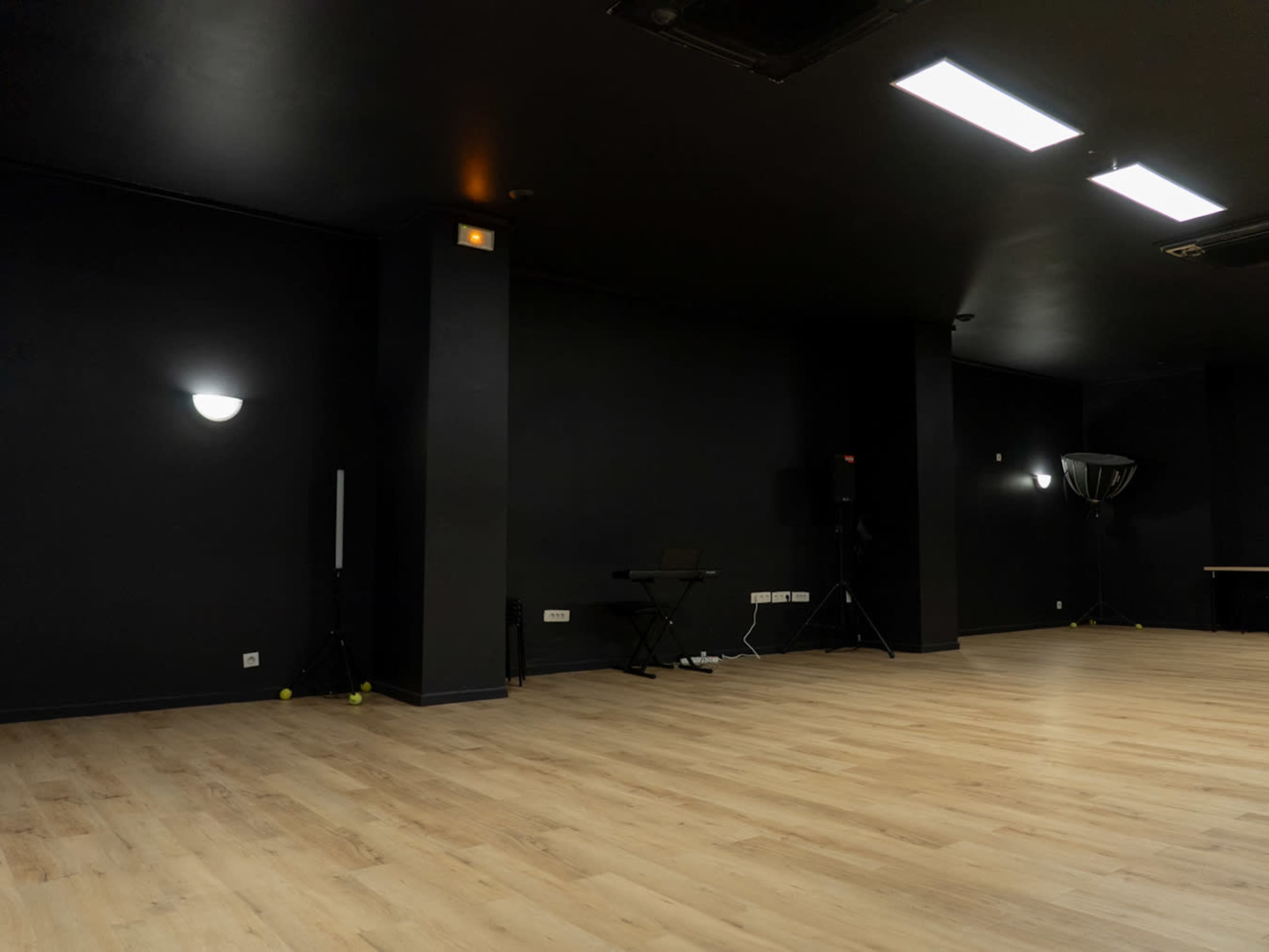The image shows a spacious room with dark walls, a wooden floor, and minimal furnishings, including a keyboard and speakers.