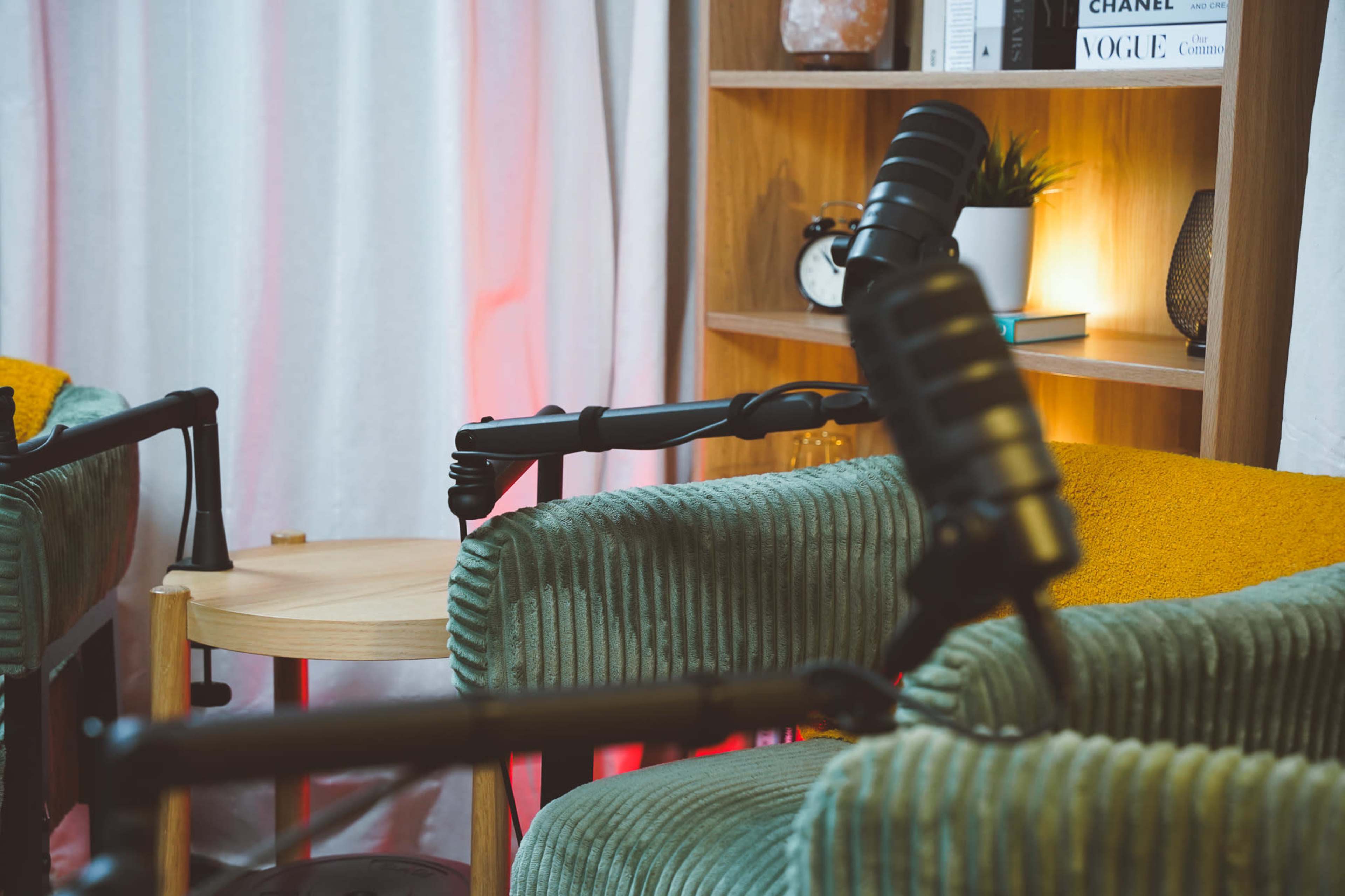 The image shows two green cushioned chairs facing each other with microphones positioned toward the center, accompanied by a small wooden table and a shelf with decorative items.