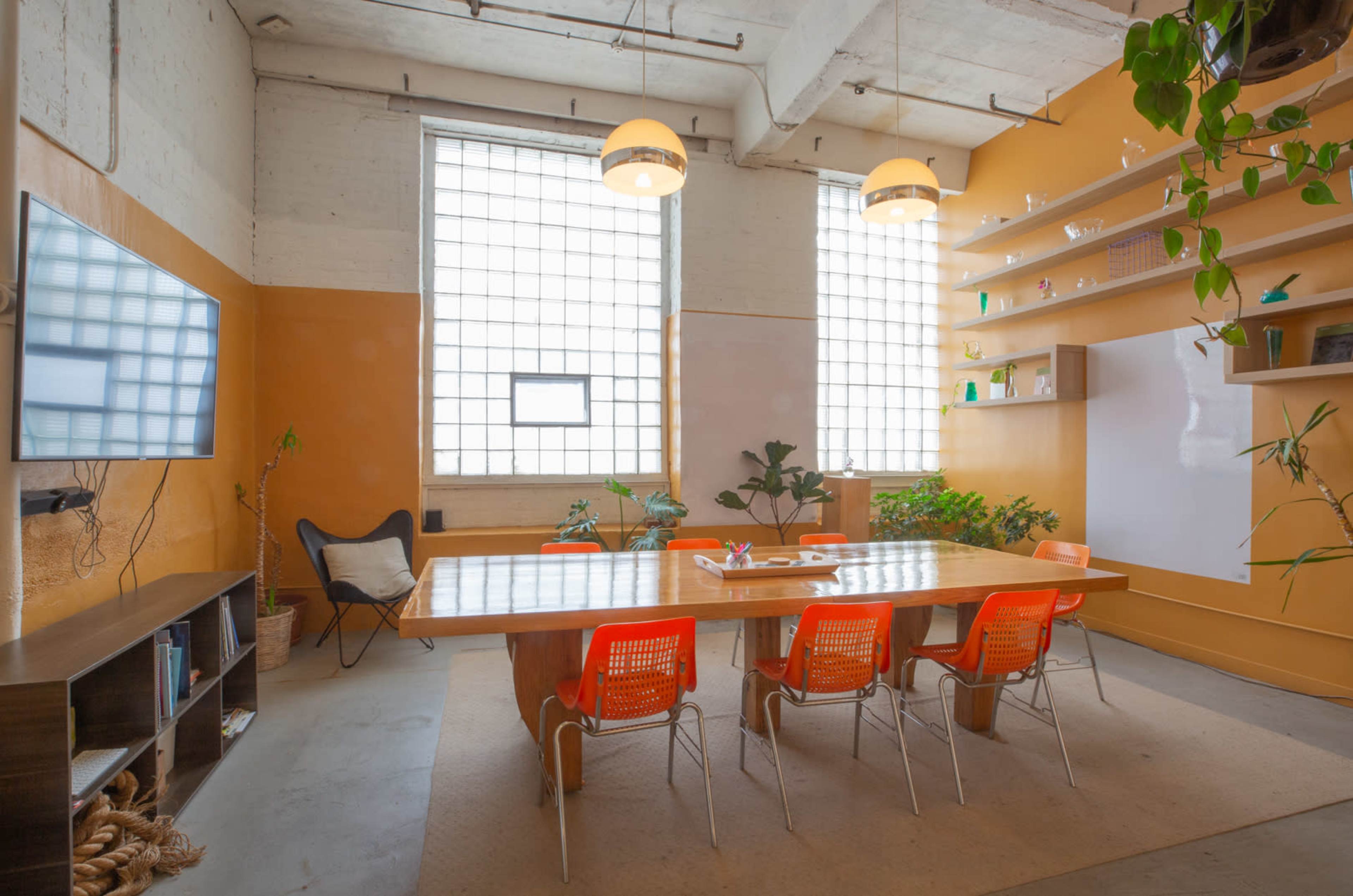 A well-lit conference room features a long wooden table surrounded by orange chairs, with large windows and plants along the walls.