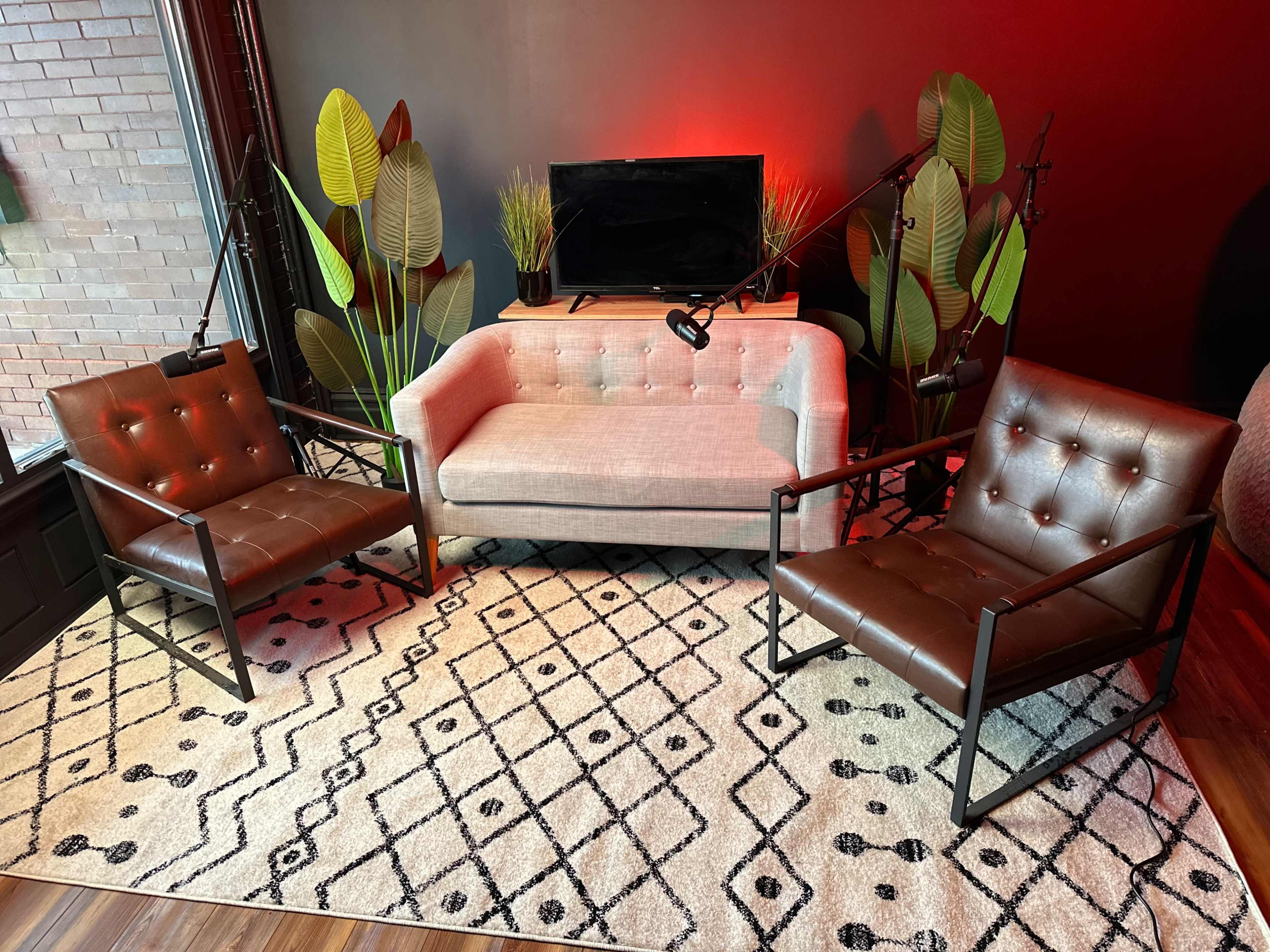 A cozy seating area with a light gray sofa and two brown chairs, set on a patterned rug under soft lighting with decorative plants in the background.