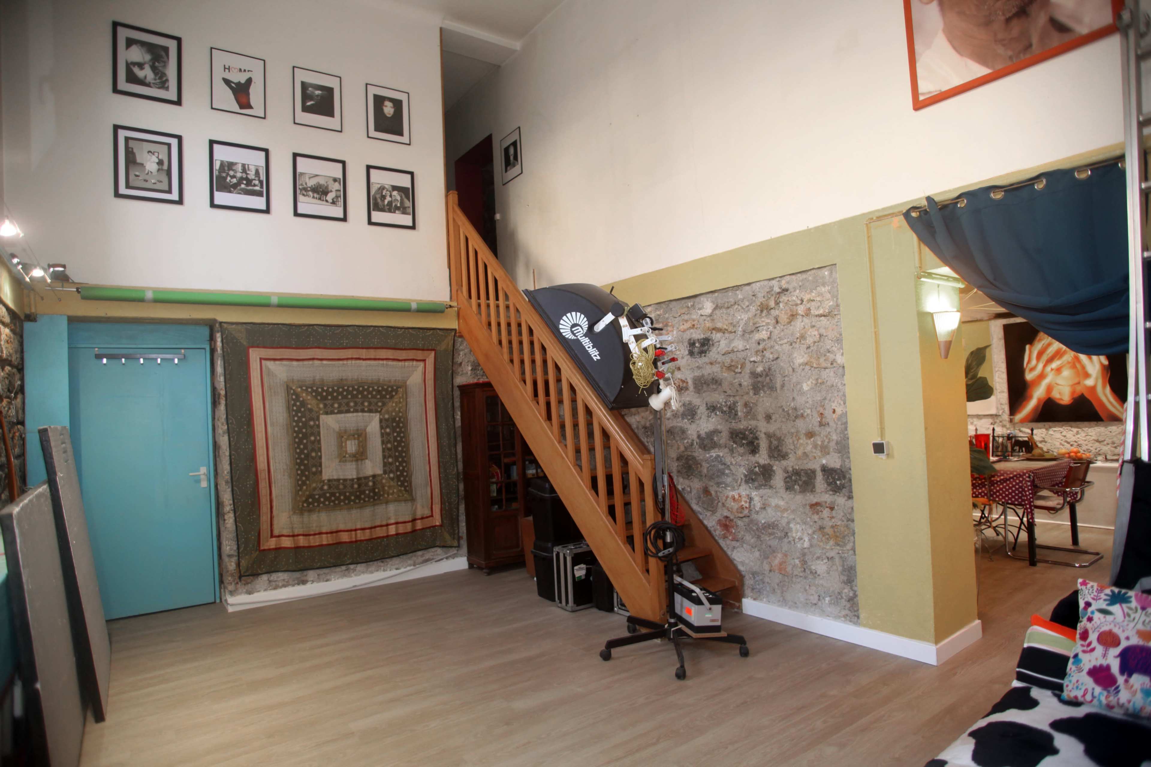 The image shows an interior space with wooden stairs leading to an upper level, framed pictures on the walls, and various decorative elements.