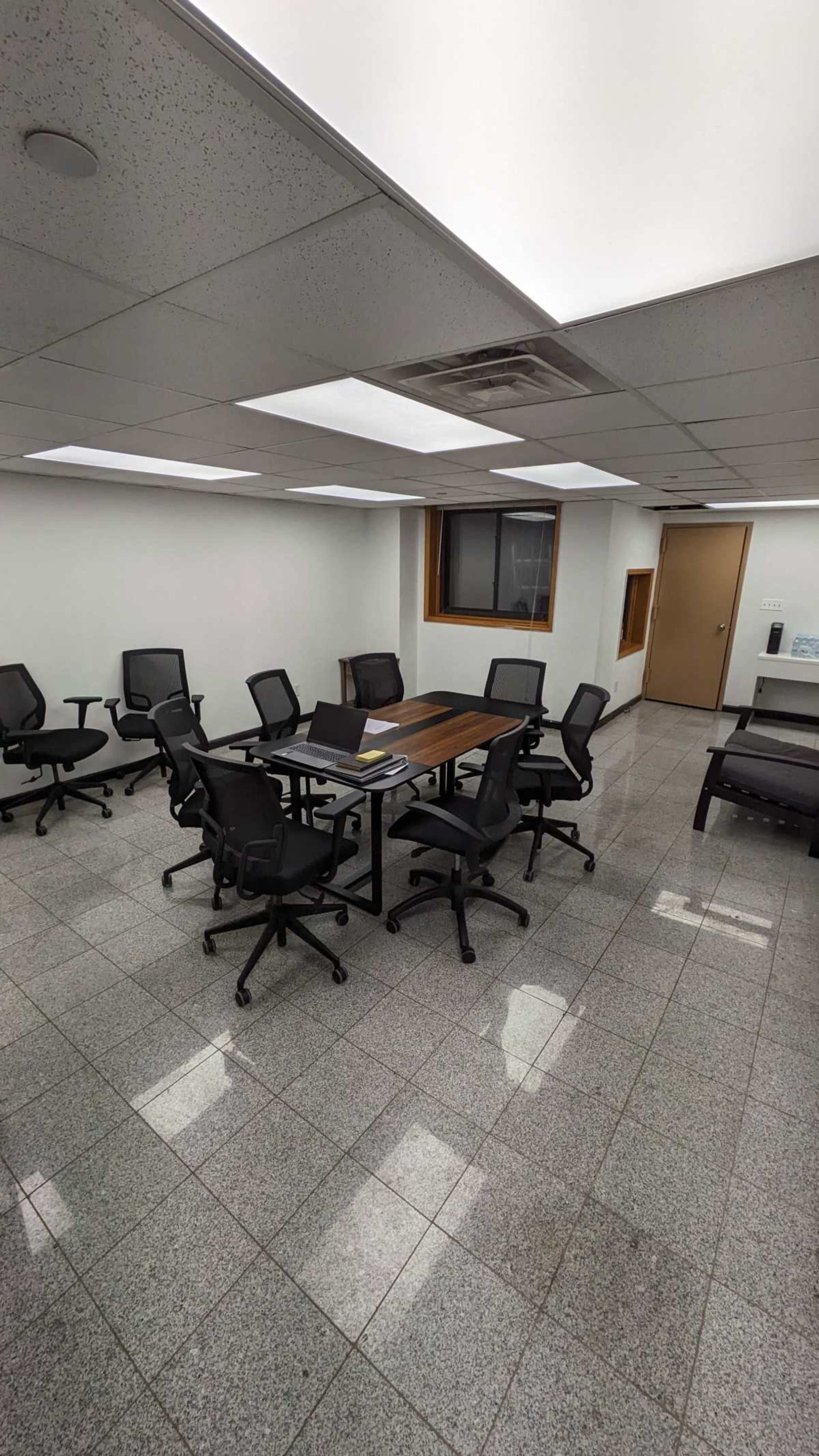 A conference room features a large rectangular table surrounded by several black ergonomic chairs on a gray tiled floor.