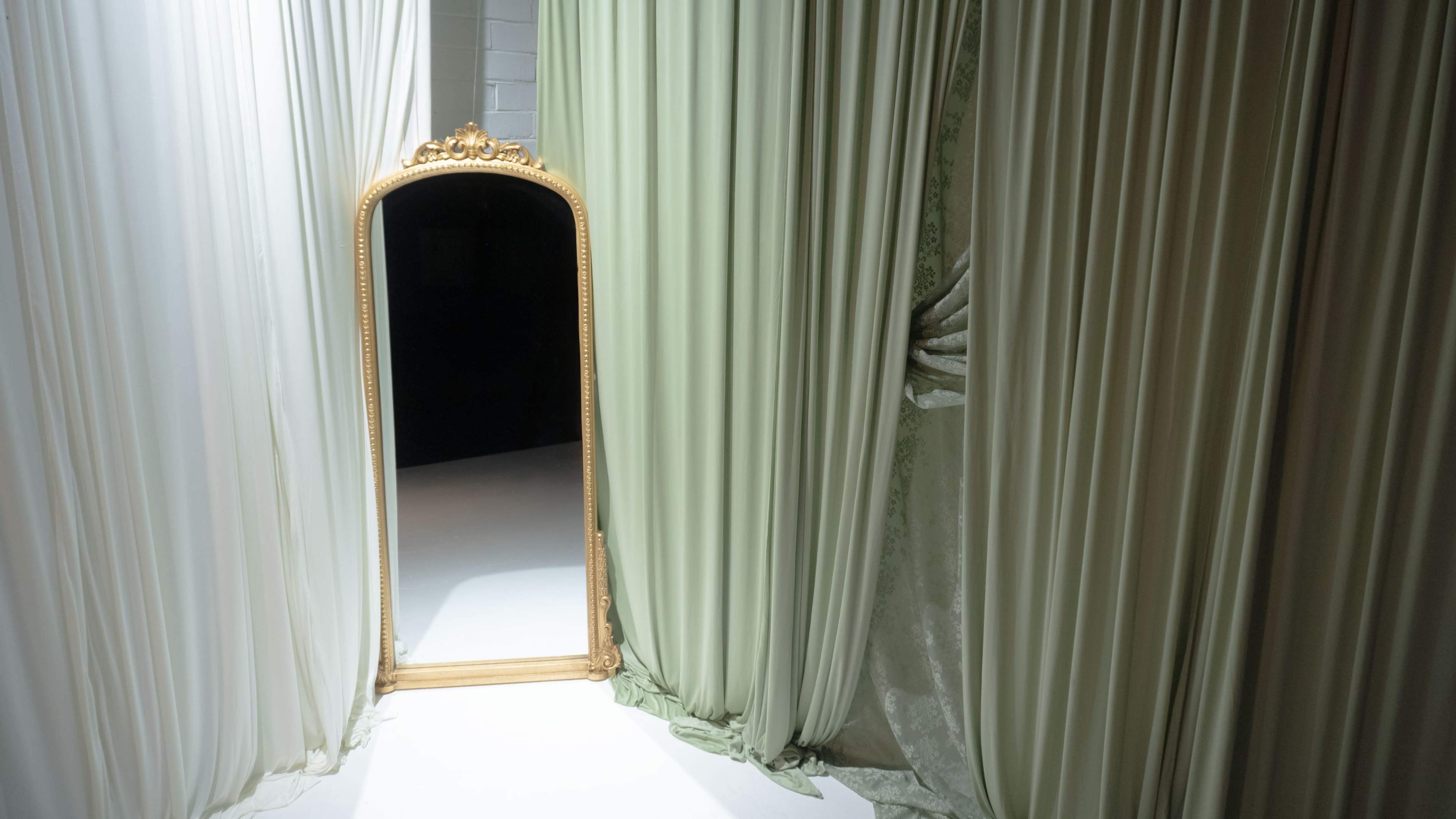 A tall, ornate mirror stands between two flowing fabric drapes in a softly lit room.