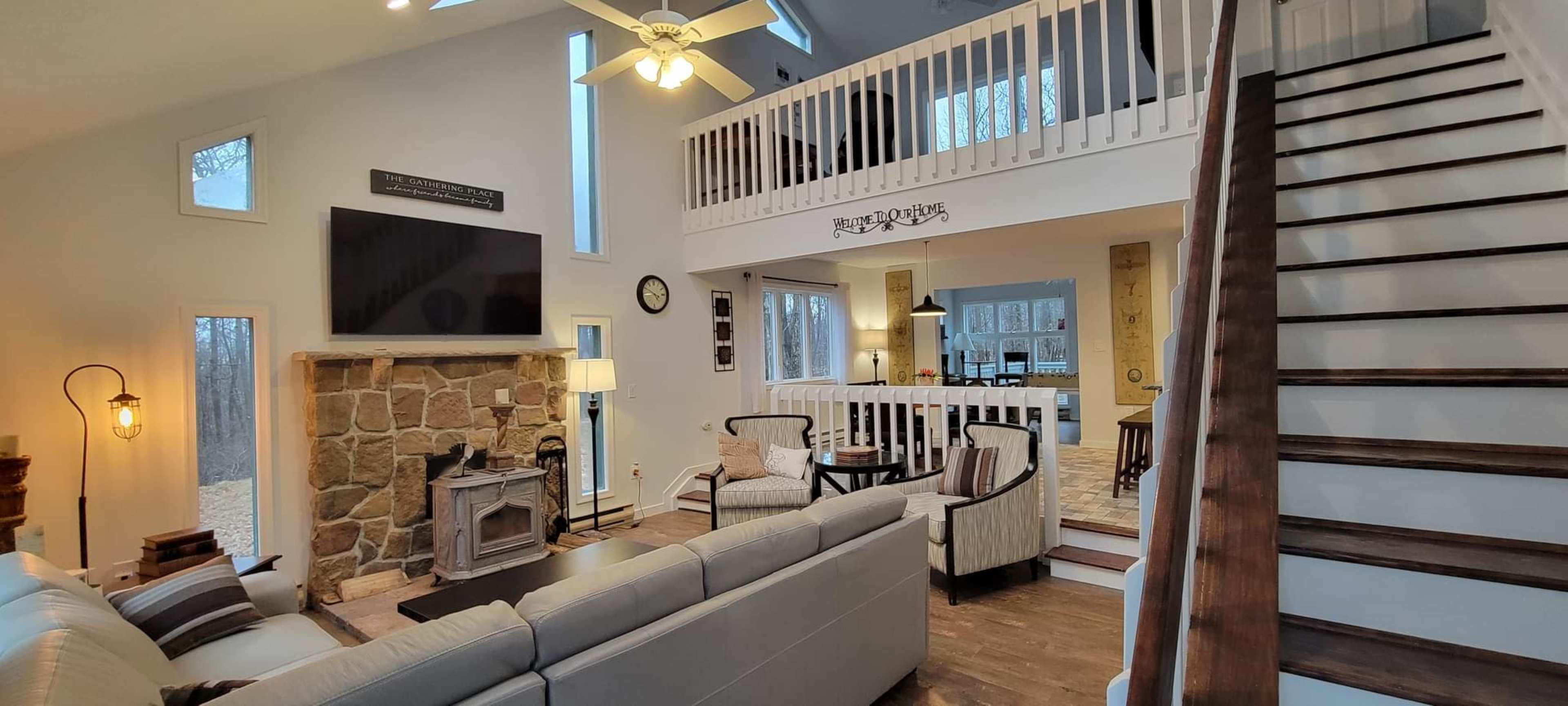 The image shows a spacious living room with a stone fireplace, large windows, and a staircase leading to a loft area.