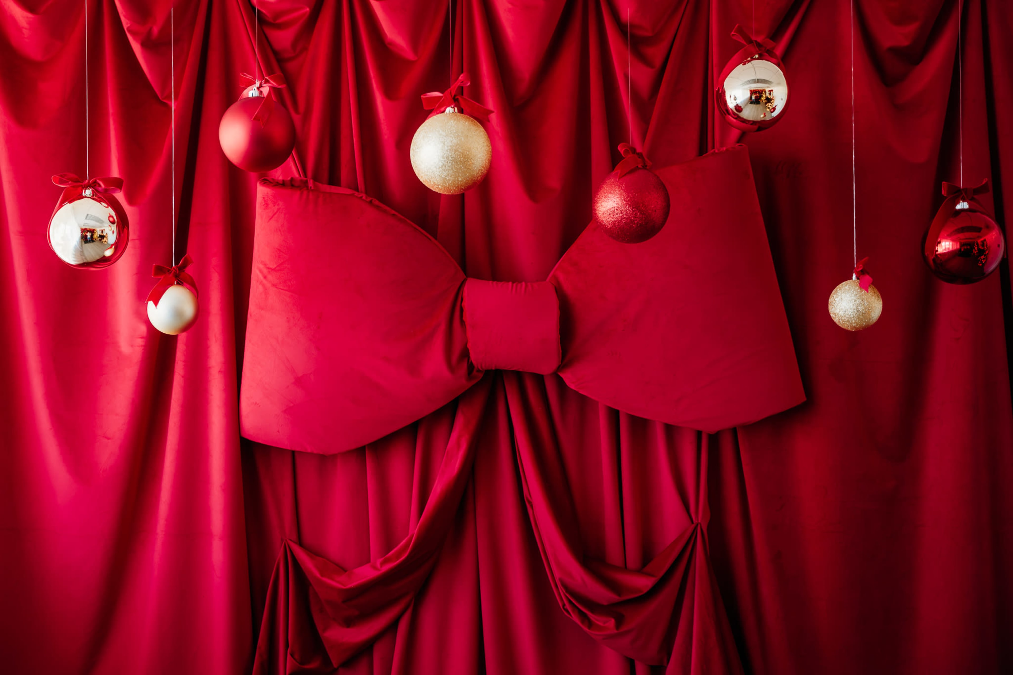 The image features a large red bow in front of a draped red fabric backdrop, adorned with festive ornaments in various colors hanging above.