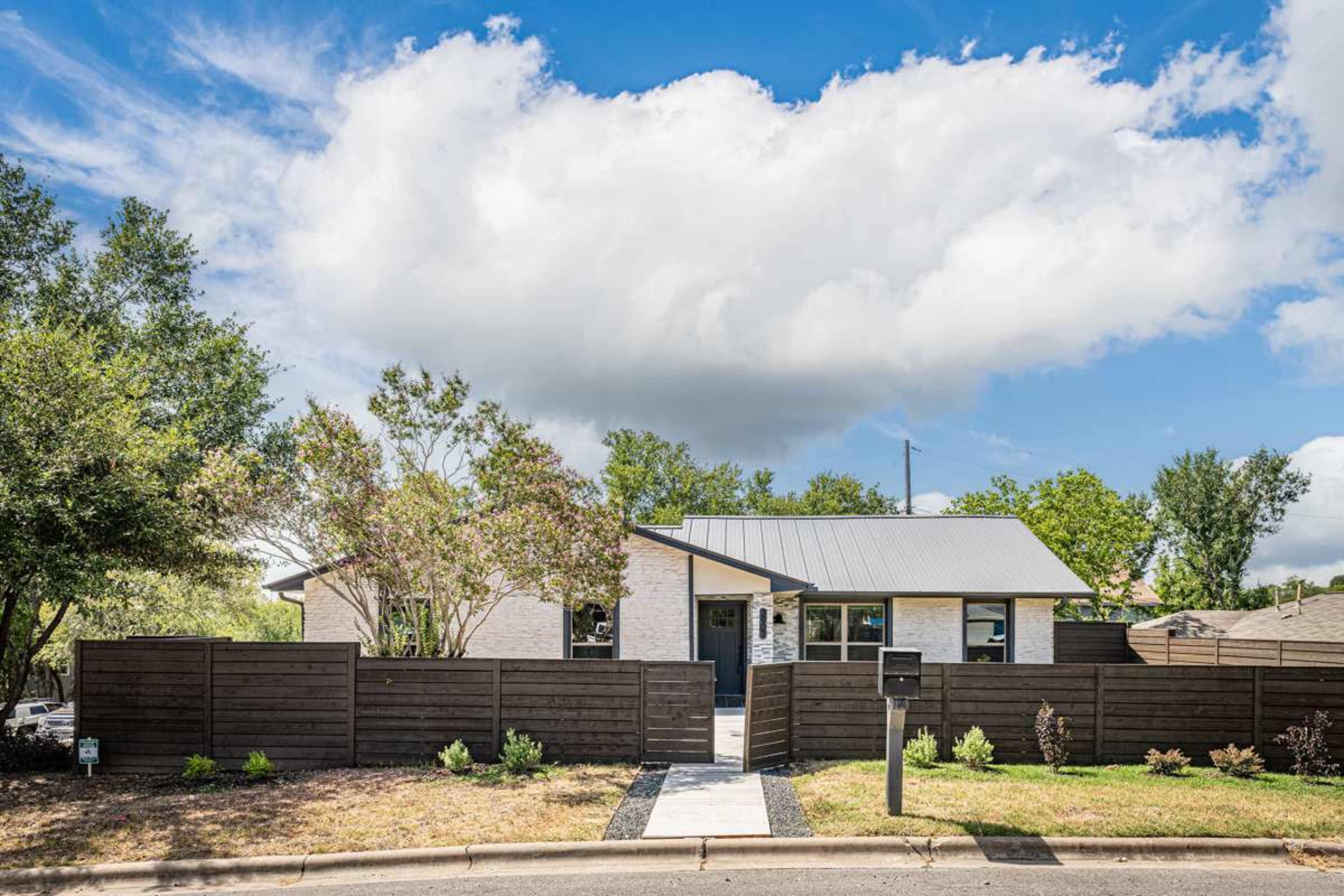 A single-story house with a metal roof is surrounded by a wooden fence and landscaped yard, set against a partly cloudy sky.