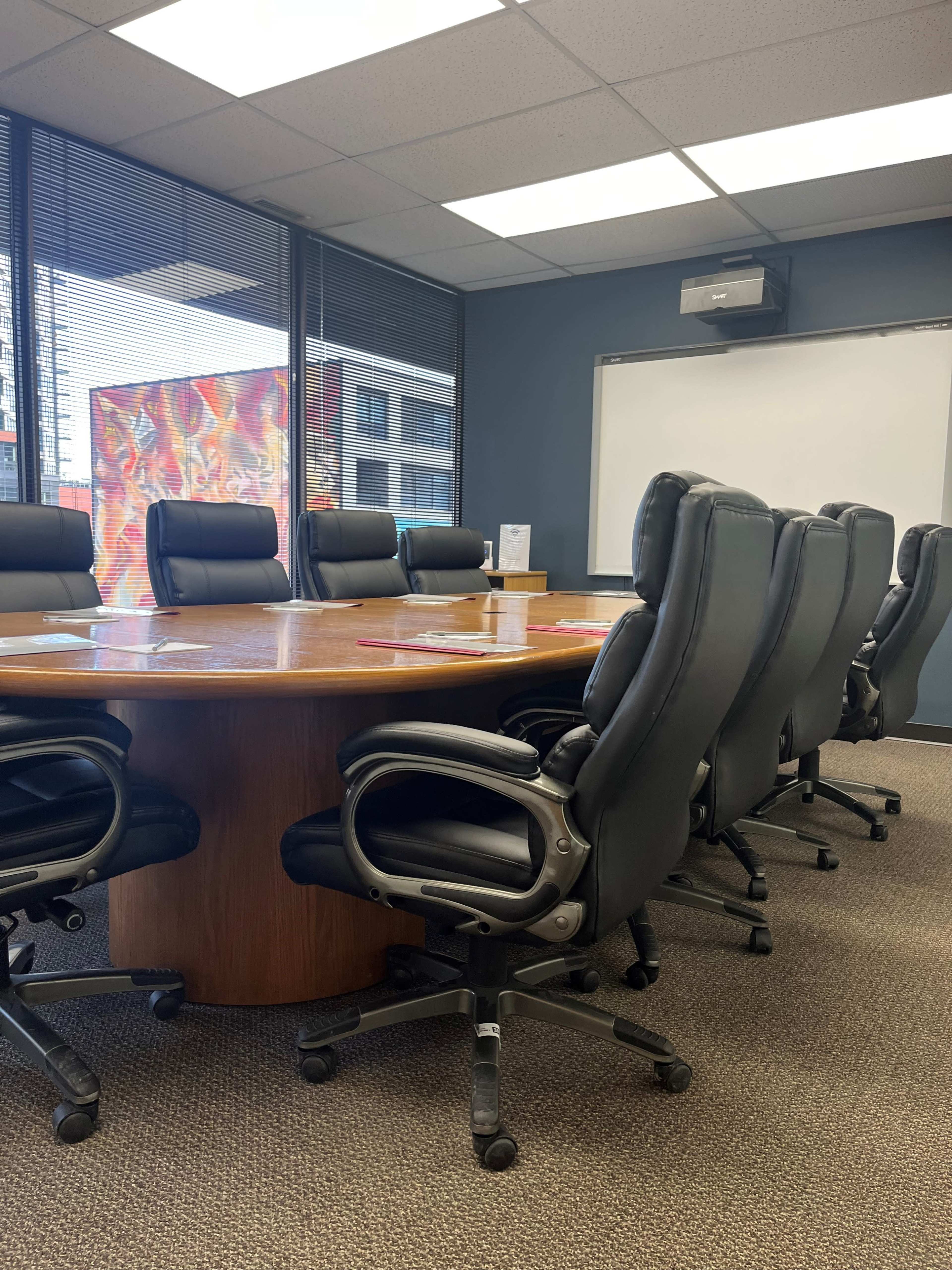 The image shows a conference room with a large wooden round table surrounded by black leather office chairs and a whiteboard on the wall.