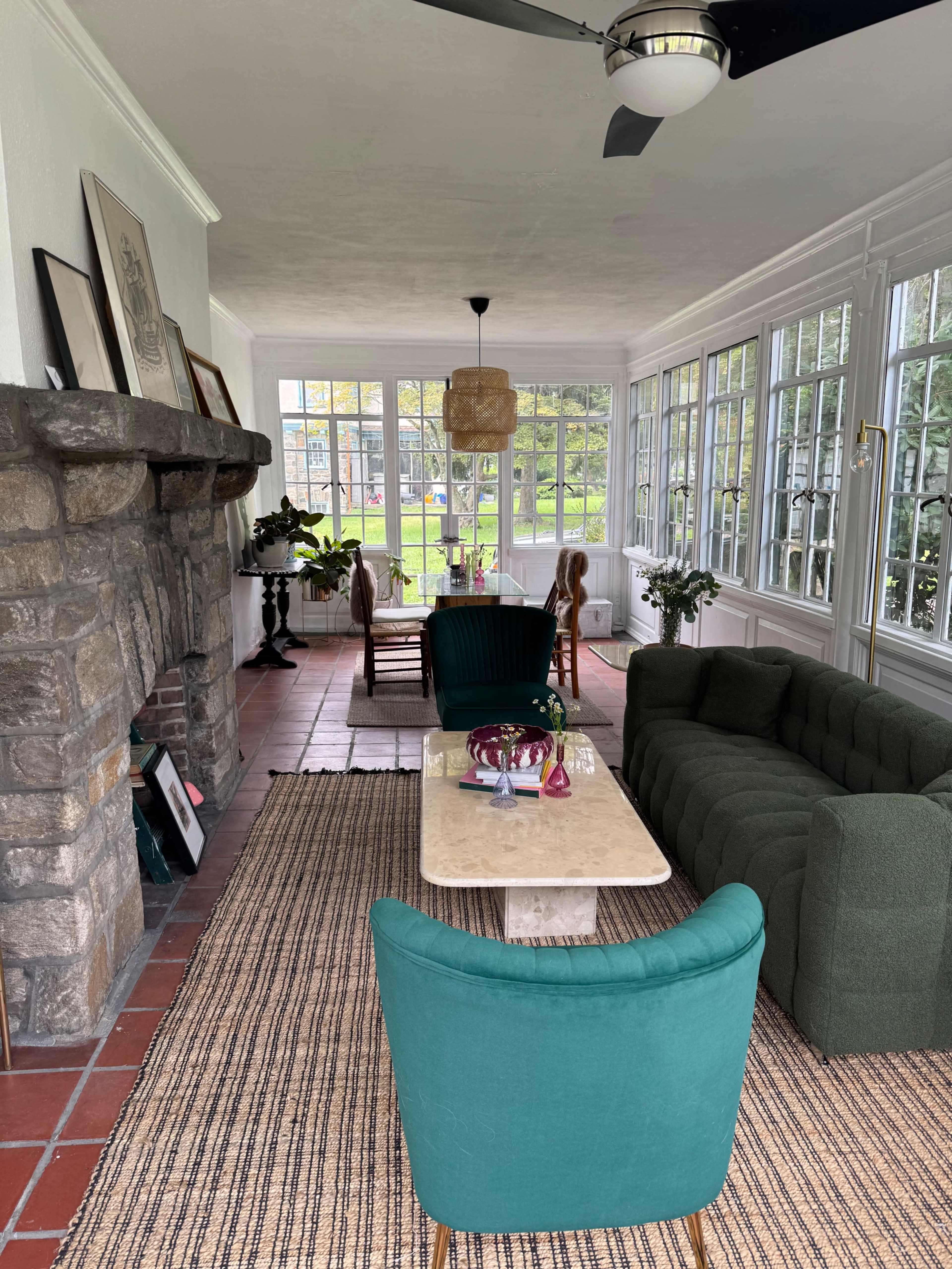 The image shows a sunlit sunroom with a stone fireplace, green furniture, and large windows overlooking a garden.