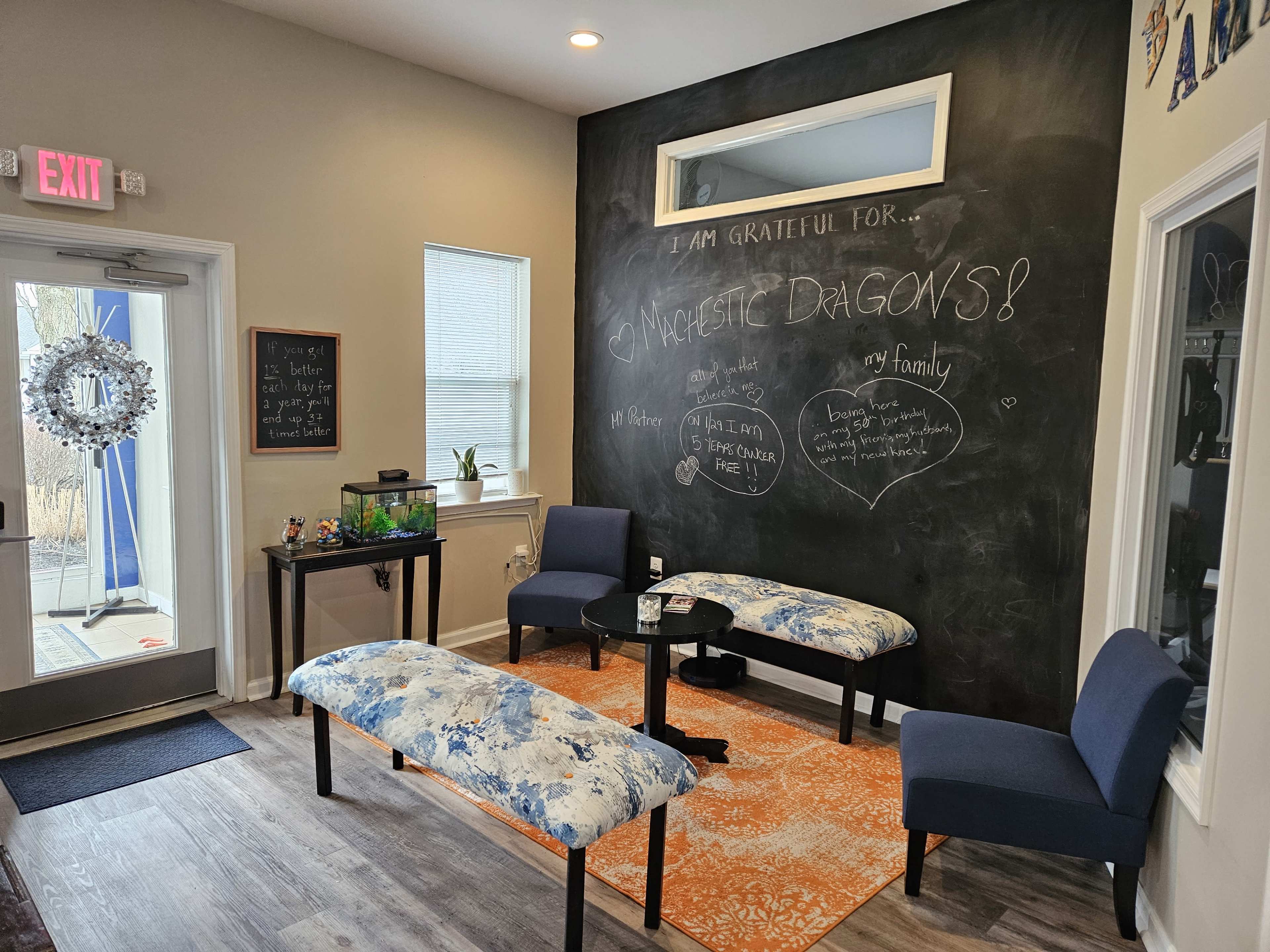 A waiting area with two blue chairs and a patterned bench, a chalkboard wall with handwritten notes, and a door leading outside.
