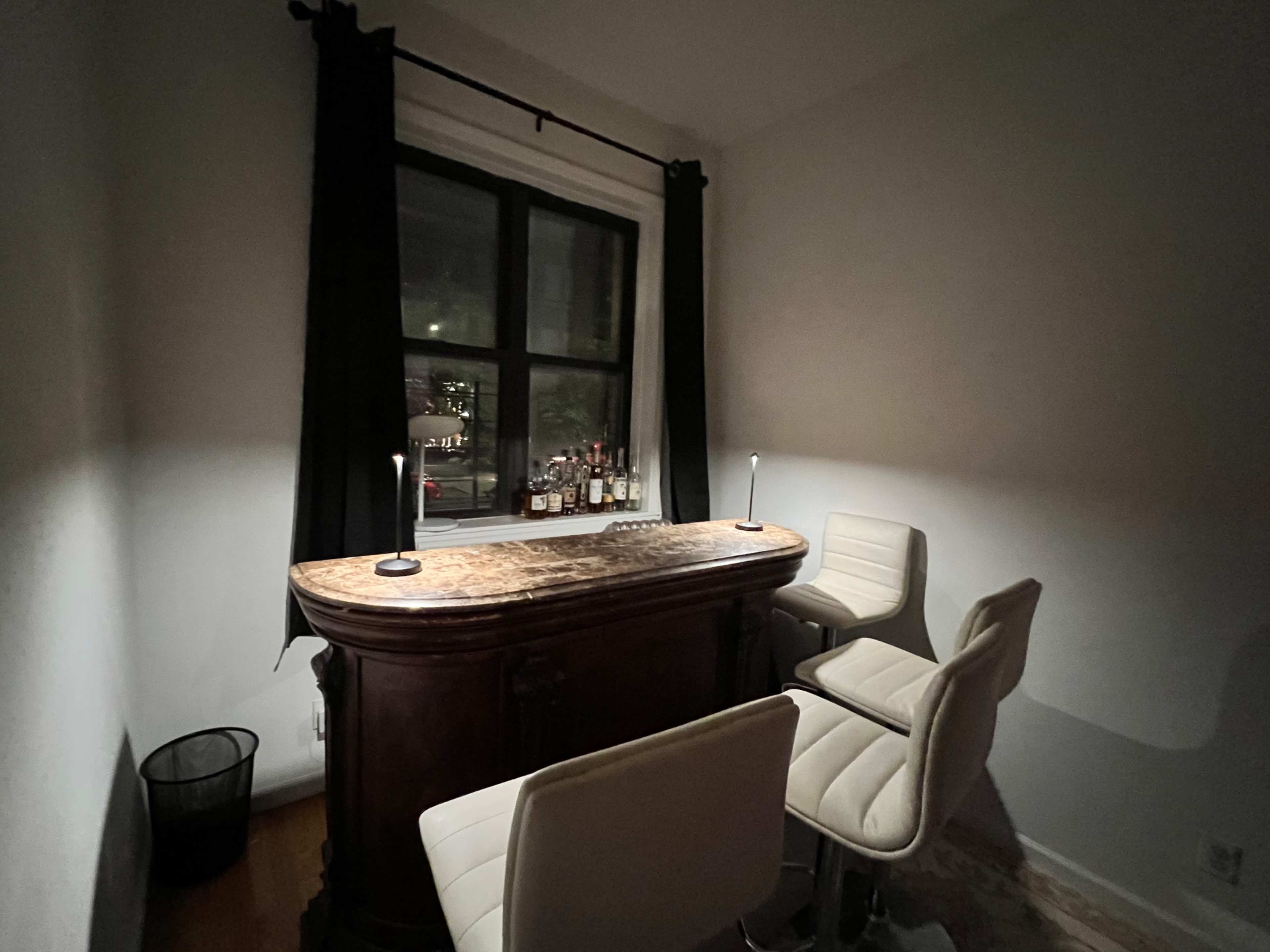 A small home bar area features a wooden bar top with two white stools, illuminated by two lamps, and a window visible in the background.