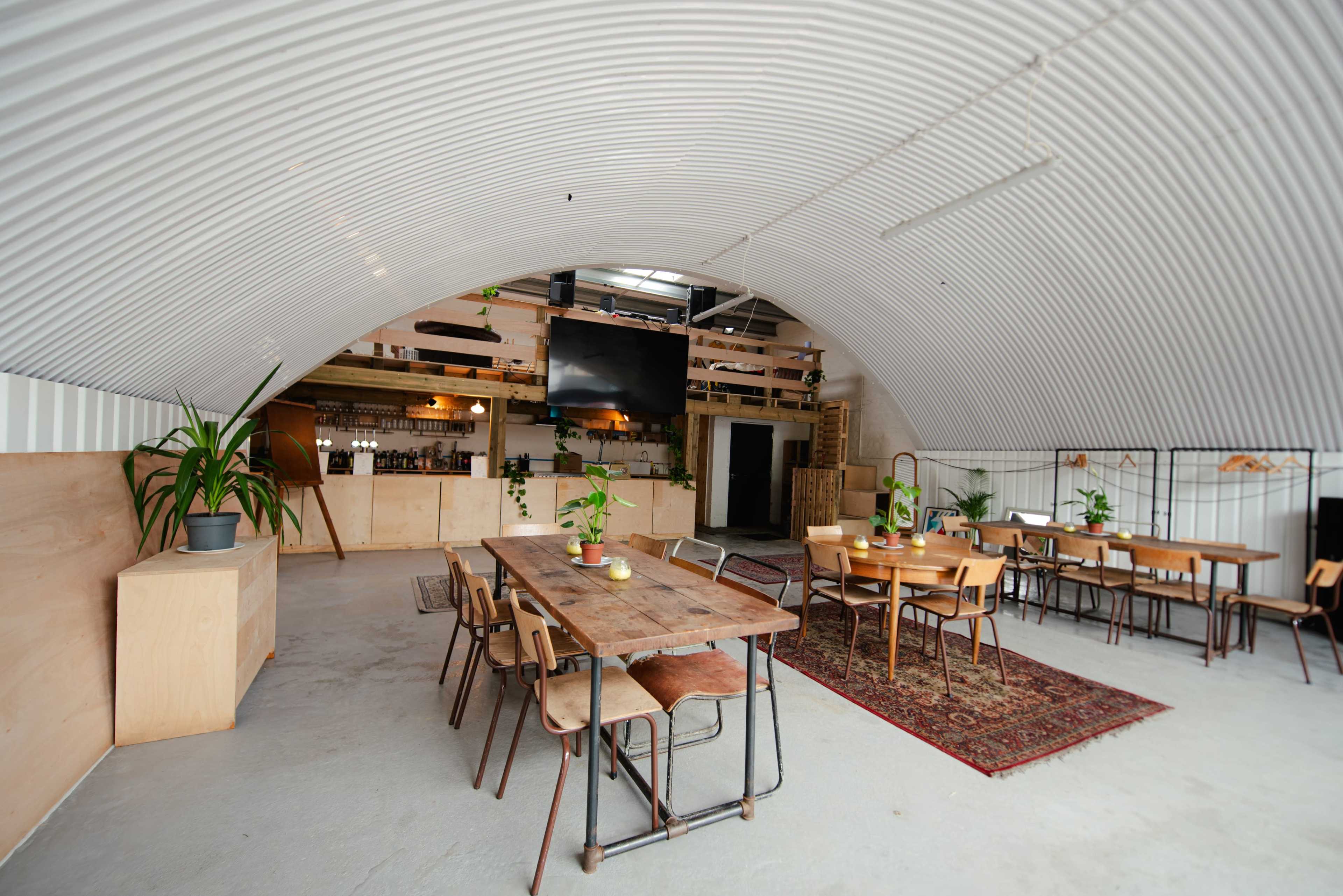 The image shows an industrial-style interior of a spacious venue with a curved metal ceiling, wooden furniture, and plants arranged on tables.