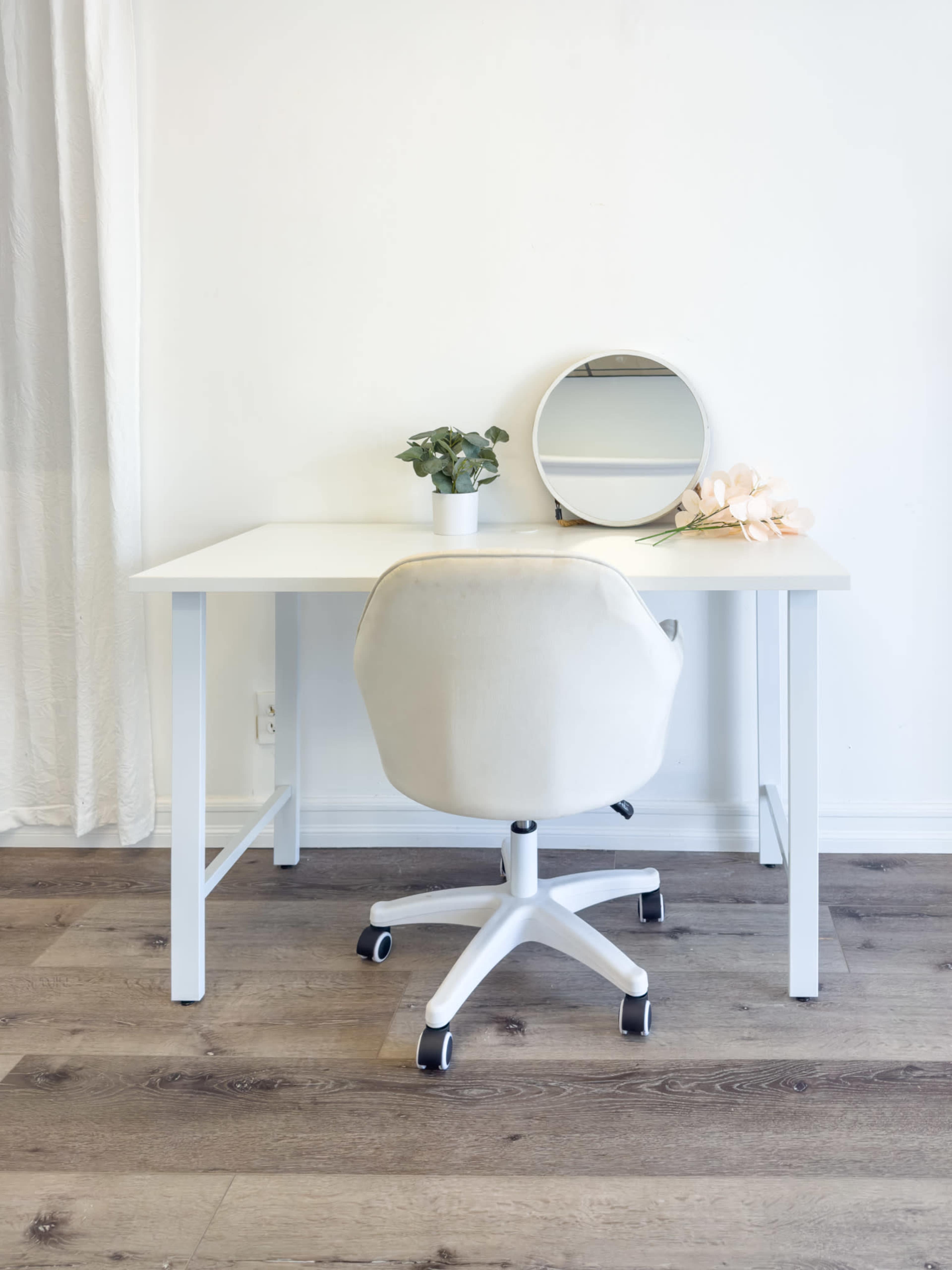A white desk with a rolling chair is positioned in front of a circular mirror, accompanied by a small potted plant and a decorative flower on the desk.