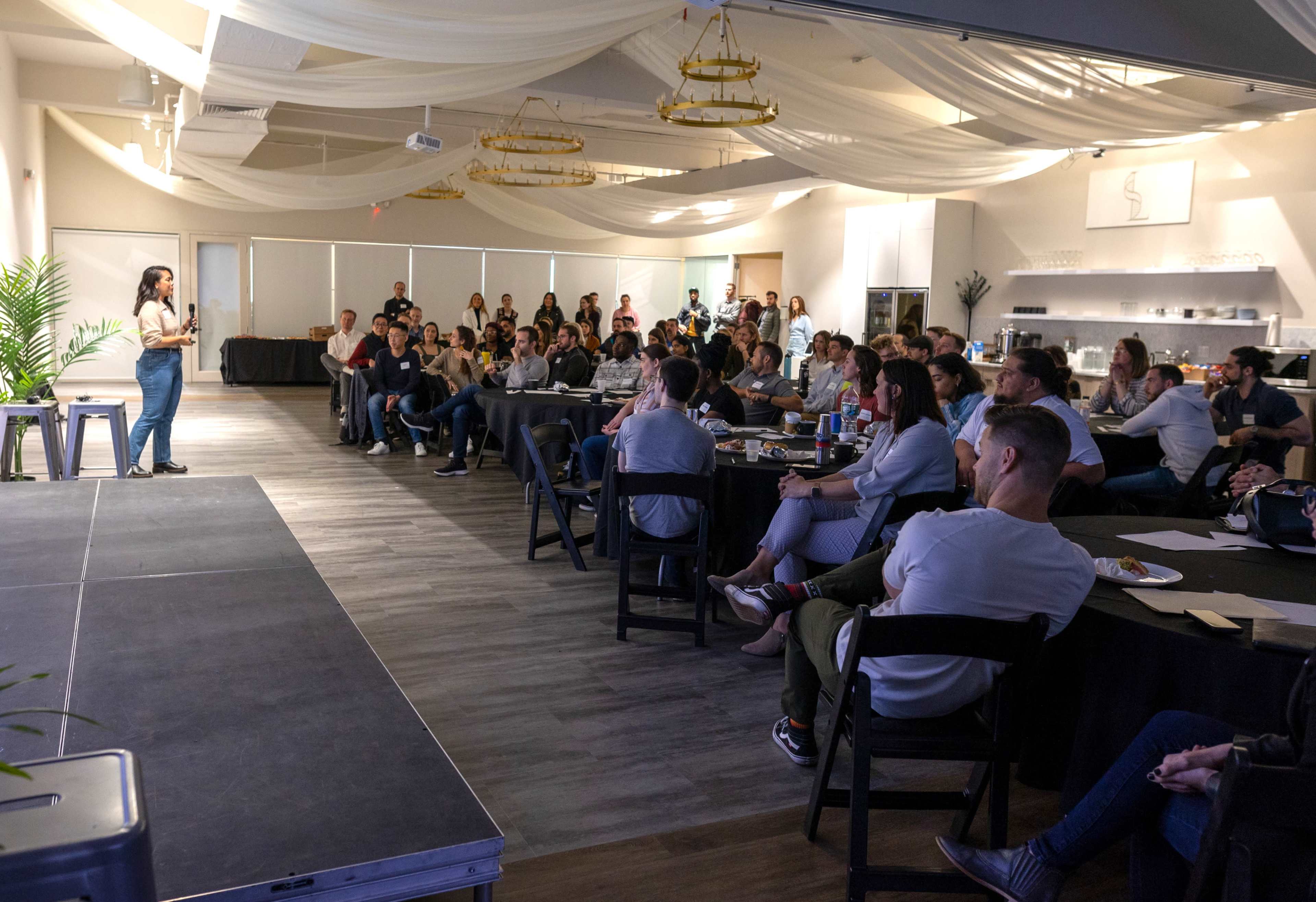 A speaker presents to an audience seated at tables in a well-lit event space with decorative elements.