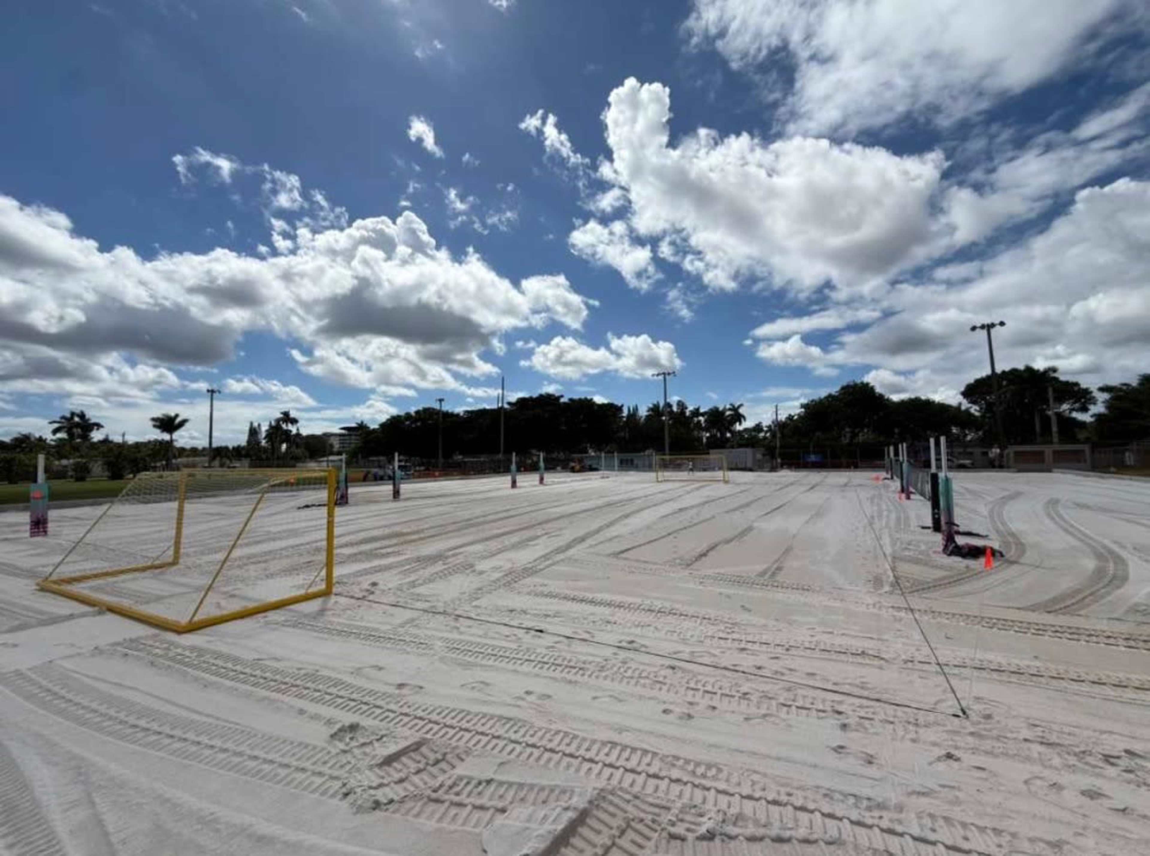 The image shows a sandy beach soccer field with two yellow goals set up under a partly cloudy sky.