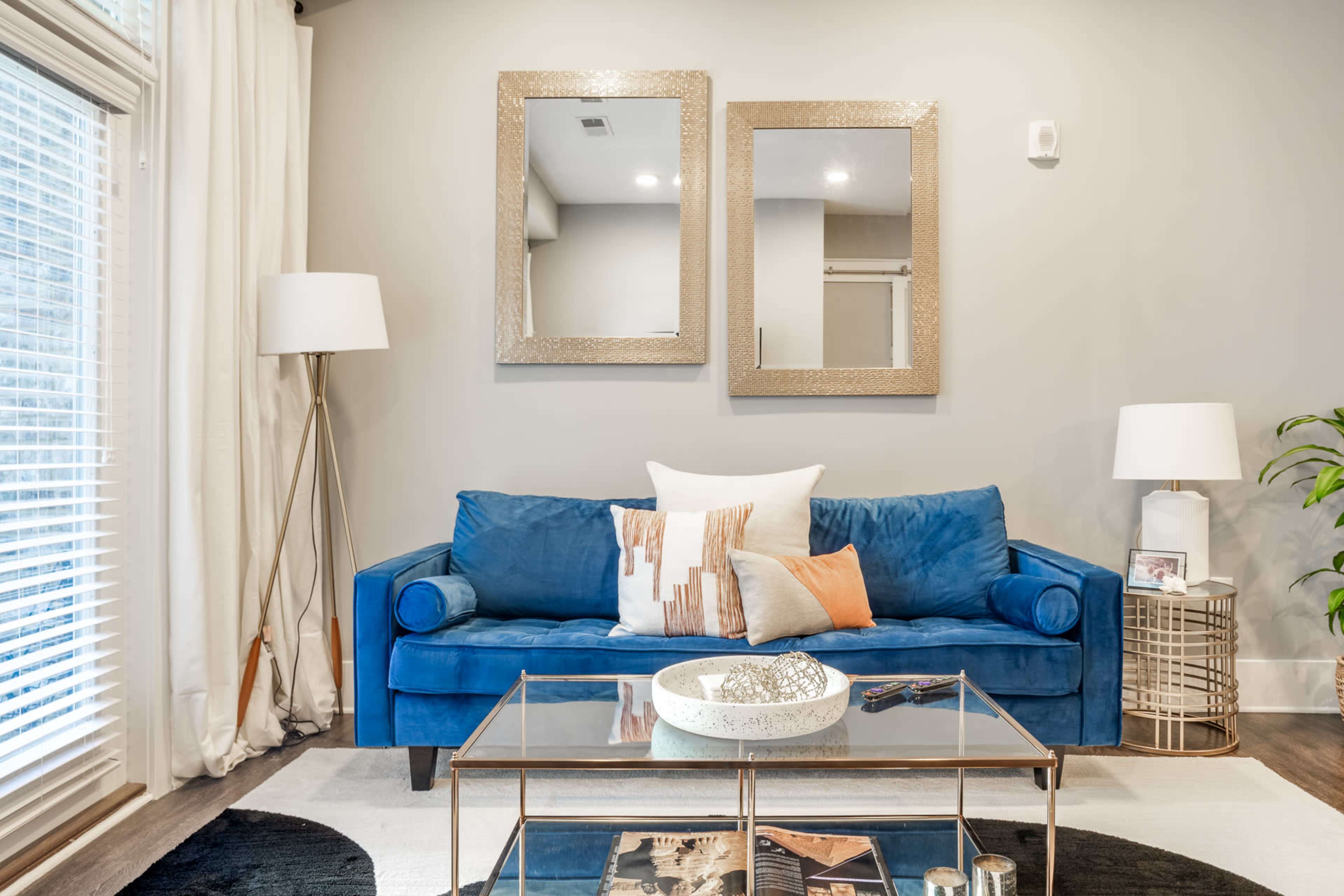 The image shows a living room featuring a blue sofa with decorative pillows, a glass coffee table, and two framed mirrors on the wall above the sofa.