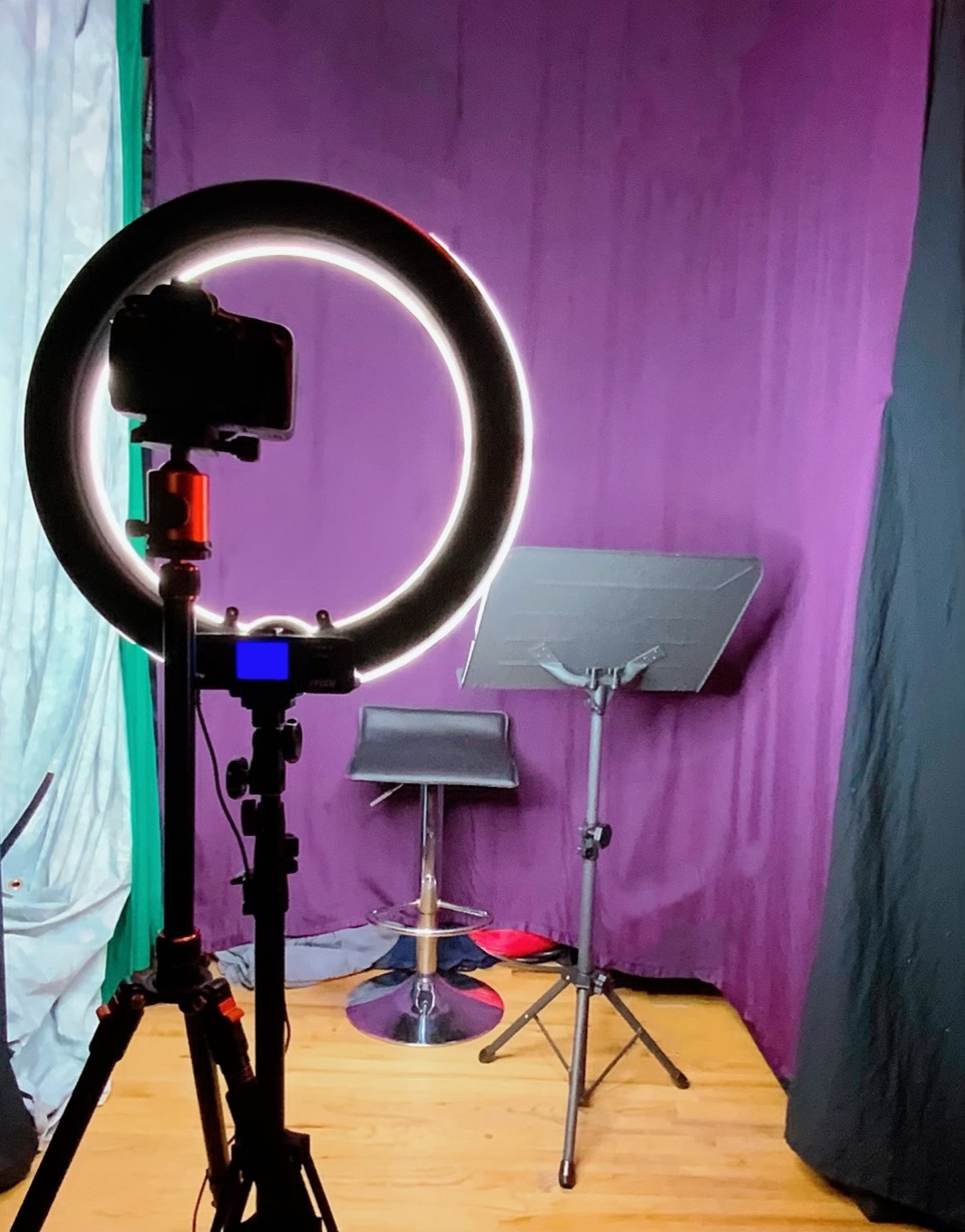 A camera mounted on a ring light is positioned in front of a purple backdrop, facing a music stand and a bar stool.