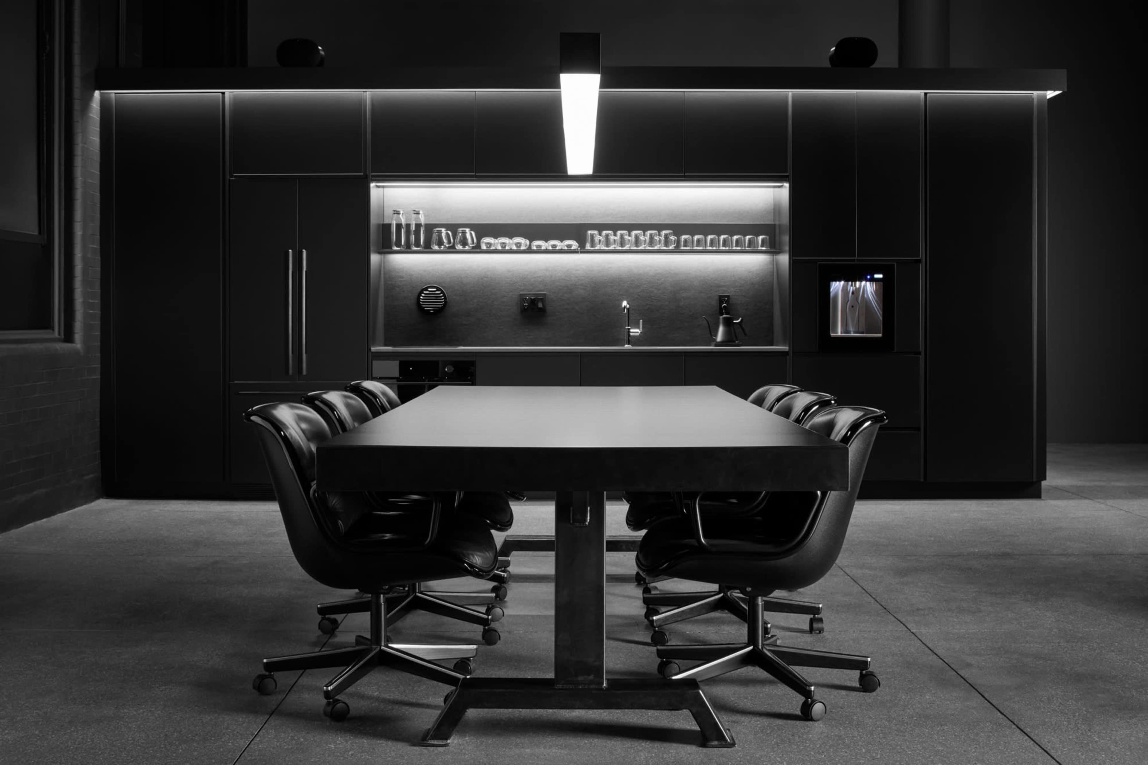 A modern kitchen features a long table surrounded by black chairs, with sleek cabinetry and minimalist appliances in the background.