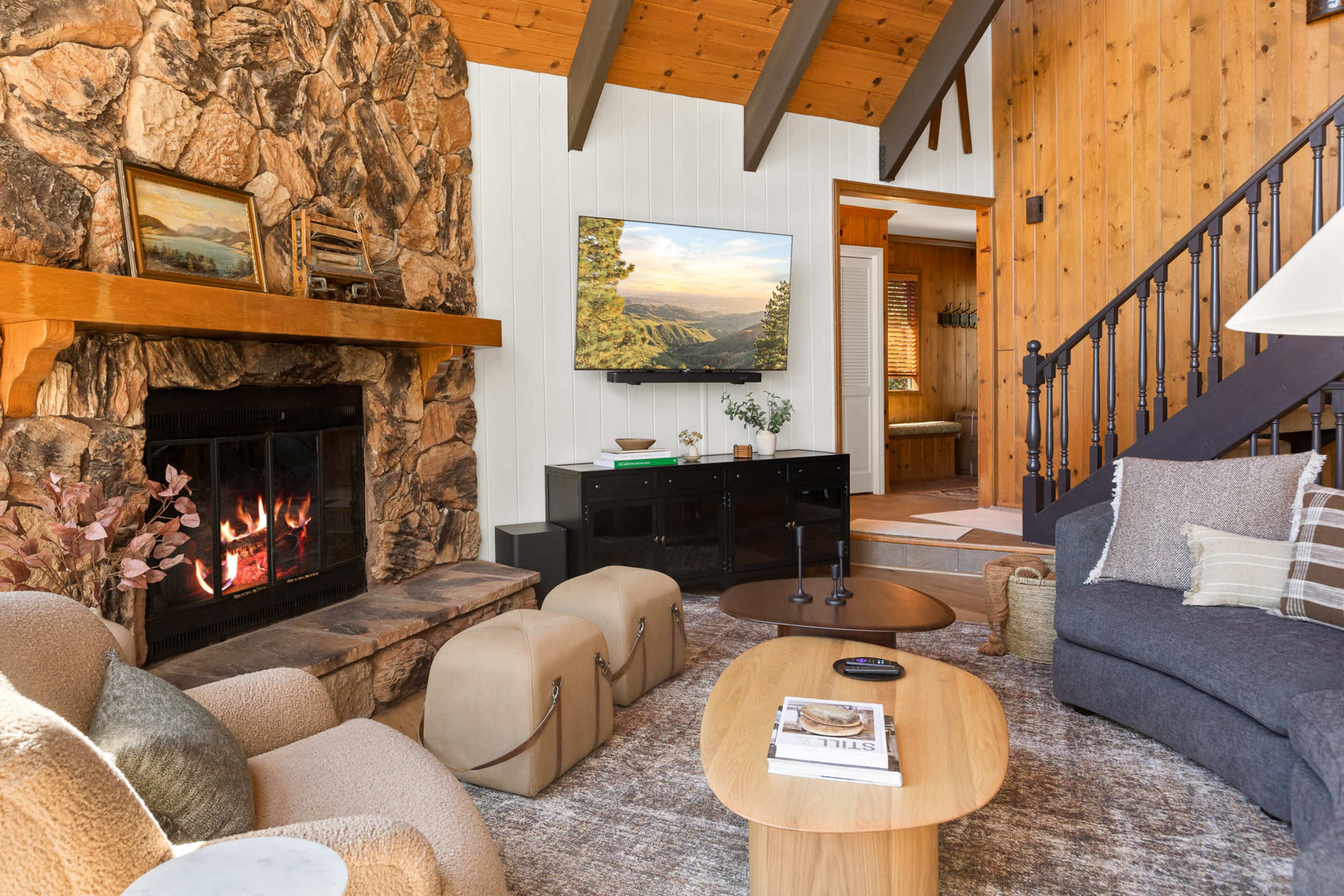A cozy living room features a stone fireplace, wooden beams, and a large television mounted on the wall, with comfortable seating and a wooden coffee table.