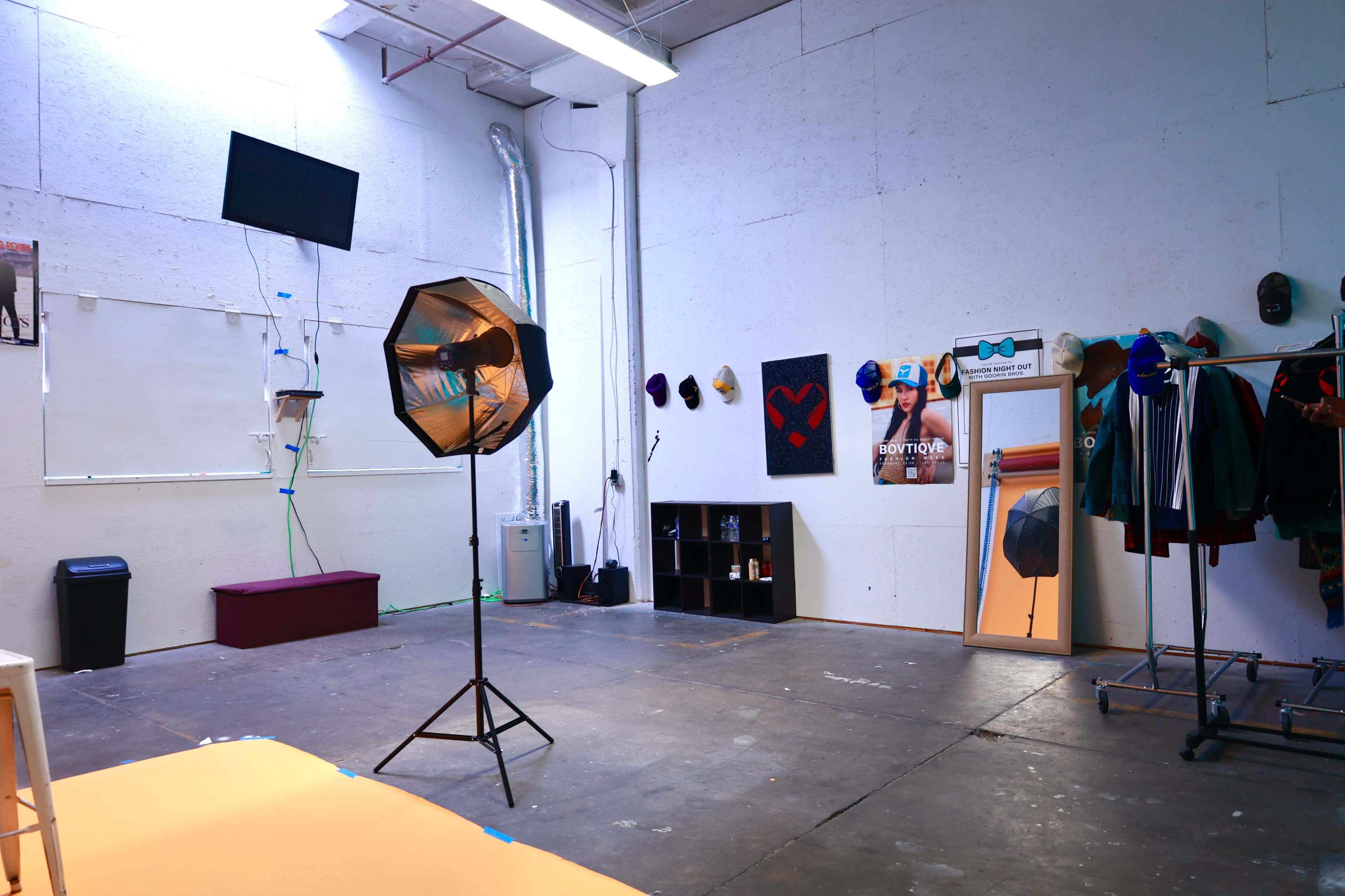 A spacious, well-lit photography studio featuring a mounted television, a large mirror, a softbox light, and various clothing items displayed on racks along the walls.