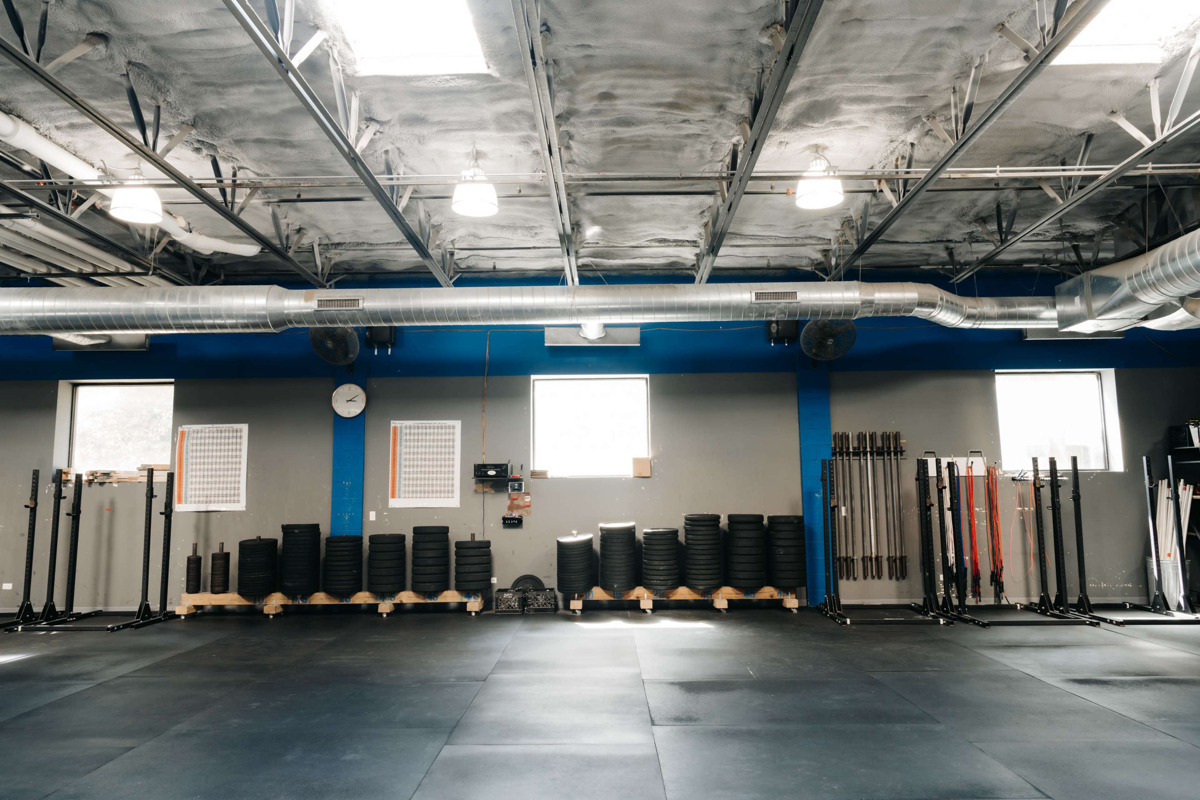The image shows a spacious gym with rubber flooring, weight plates stacked against the wall, and several barbells positioned nearby.
