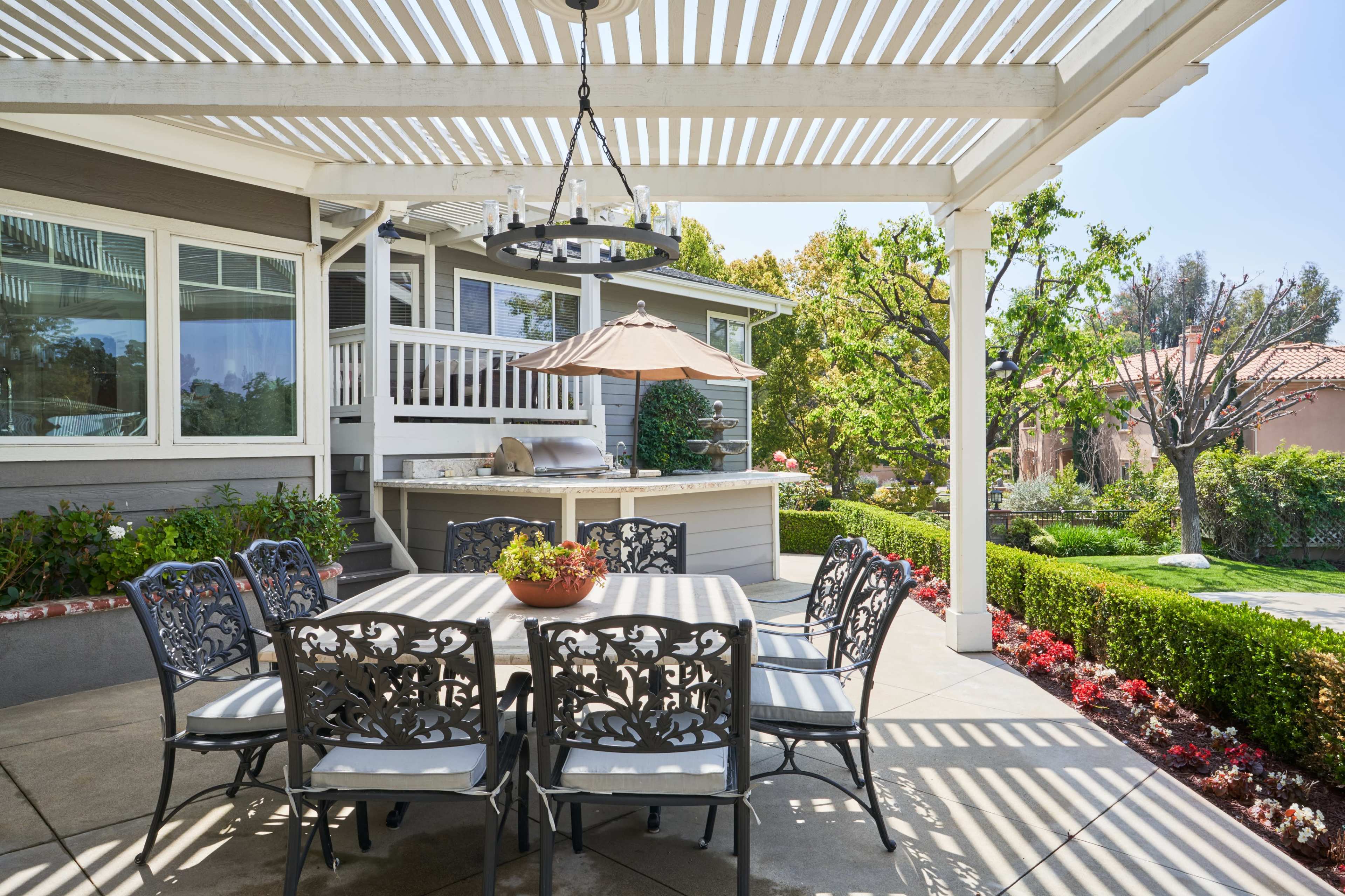 A patio area features a table with six chairs under a pergola, surrounded by well-maintained gardens and a bar space nearby.