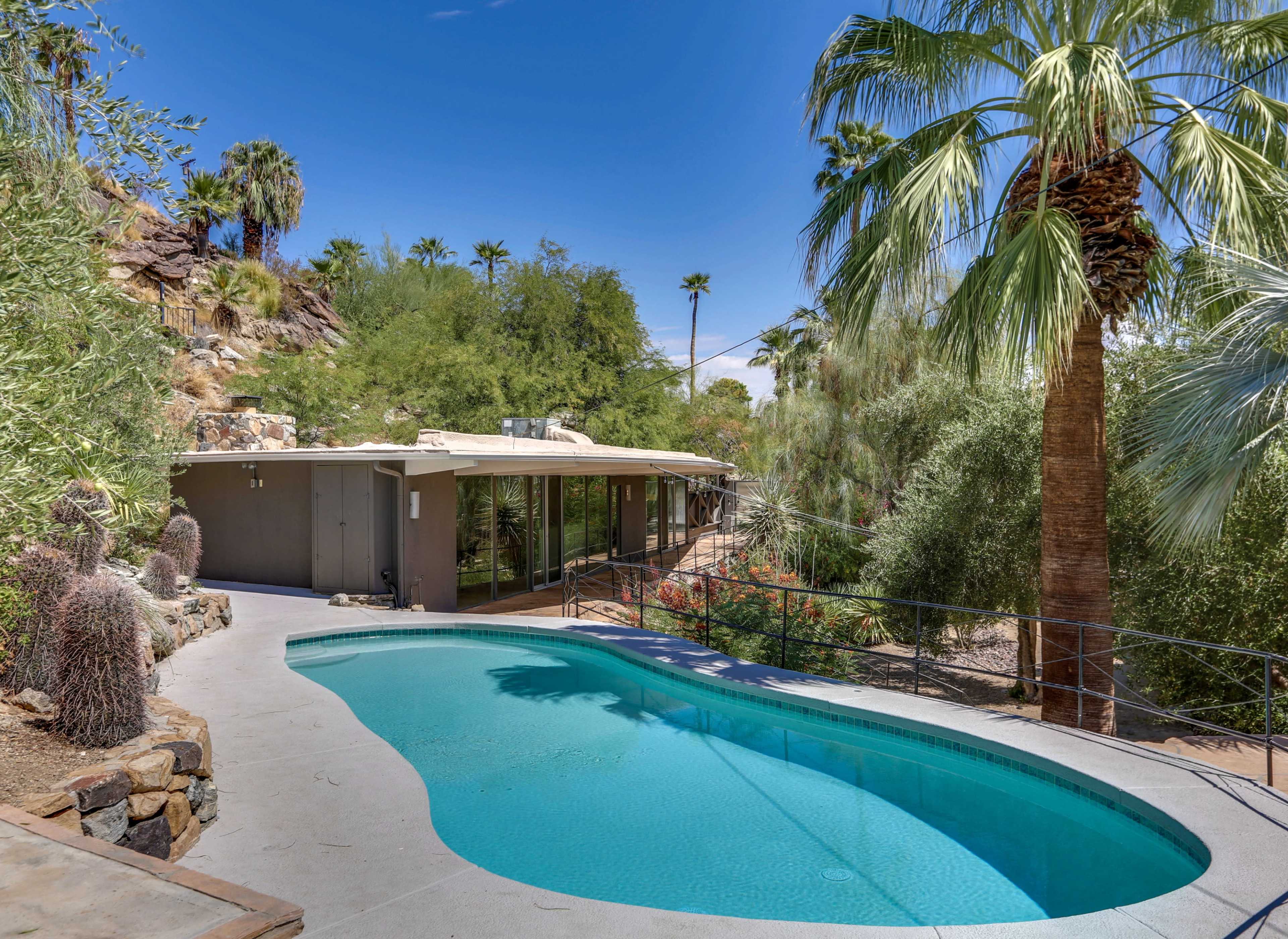 A modern home with large windows overlooks a swimming pool surrounded by palm trees and desert landscaping.