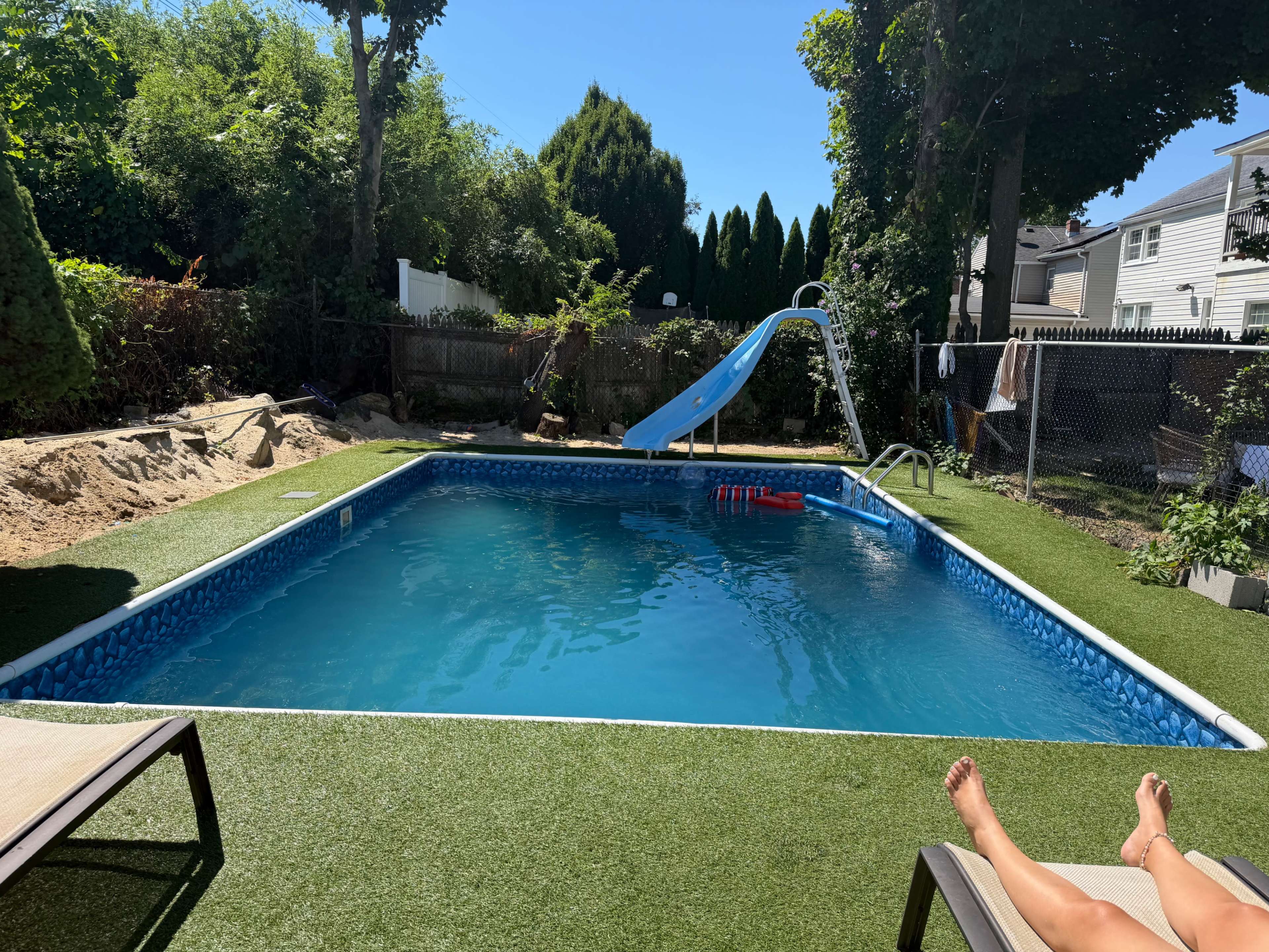 The image shows a rectangular swimming pool with a blue slide, surrounded by green grass and trees, and a lounge chair in the foreground.