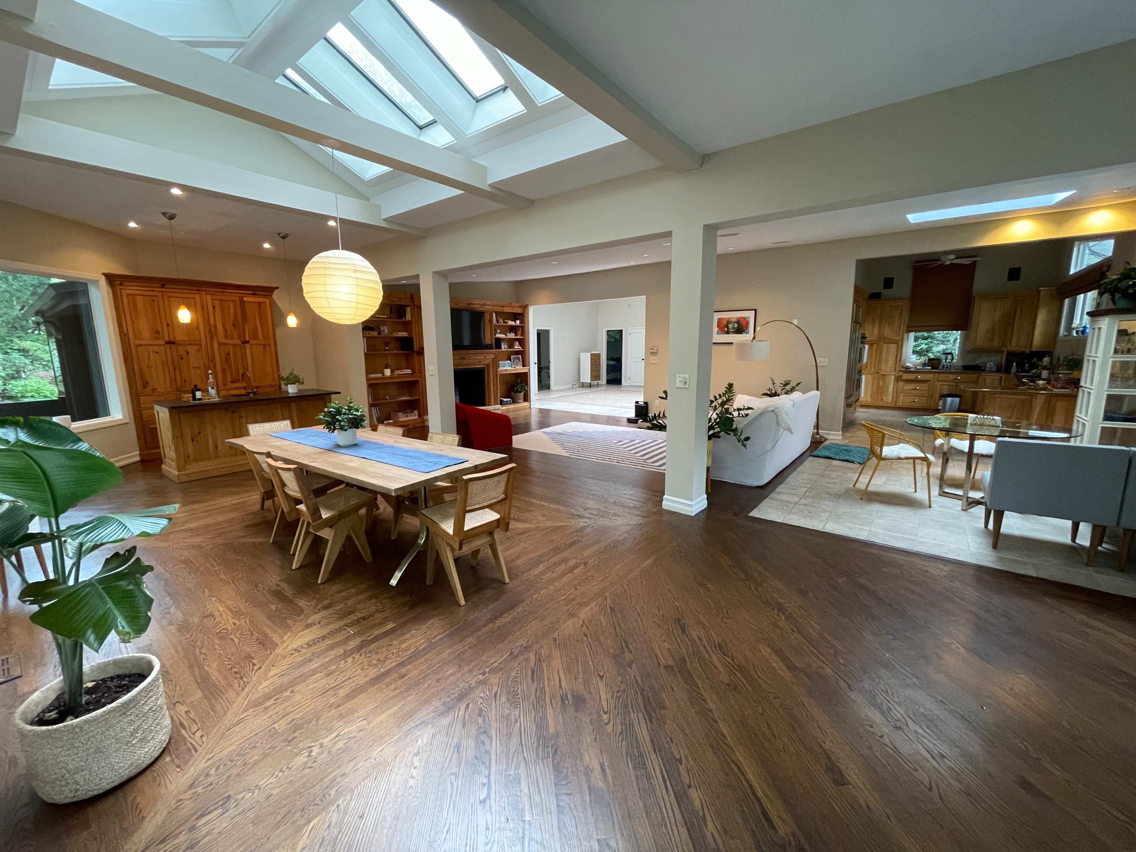 An open-concept living space with a dining area featuring a wooden table, a kitchen in the background, and large windows allowing natural light to enter.