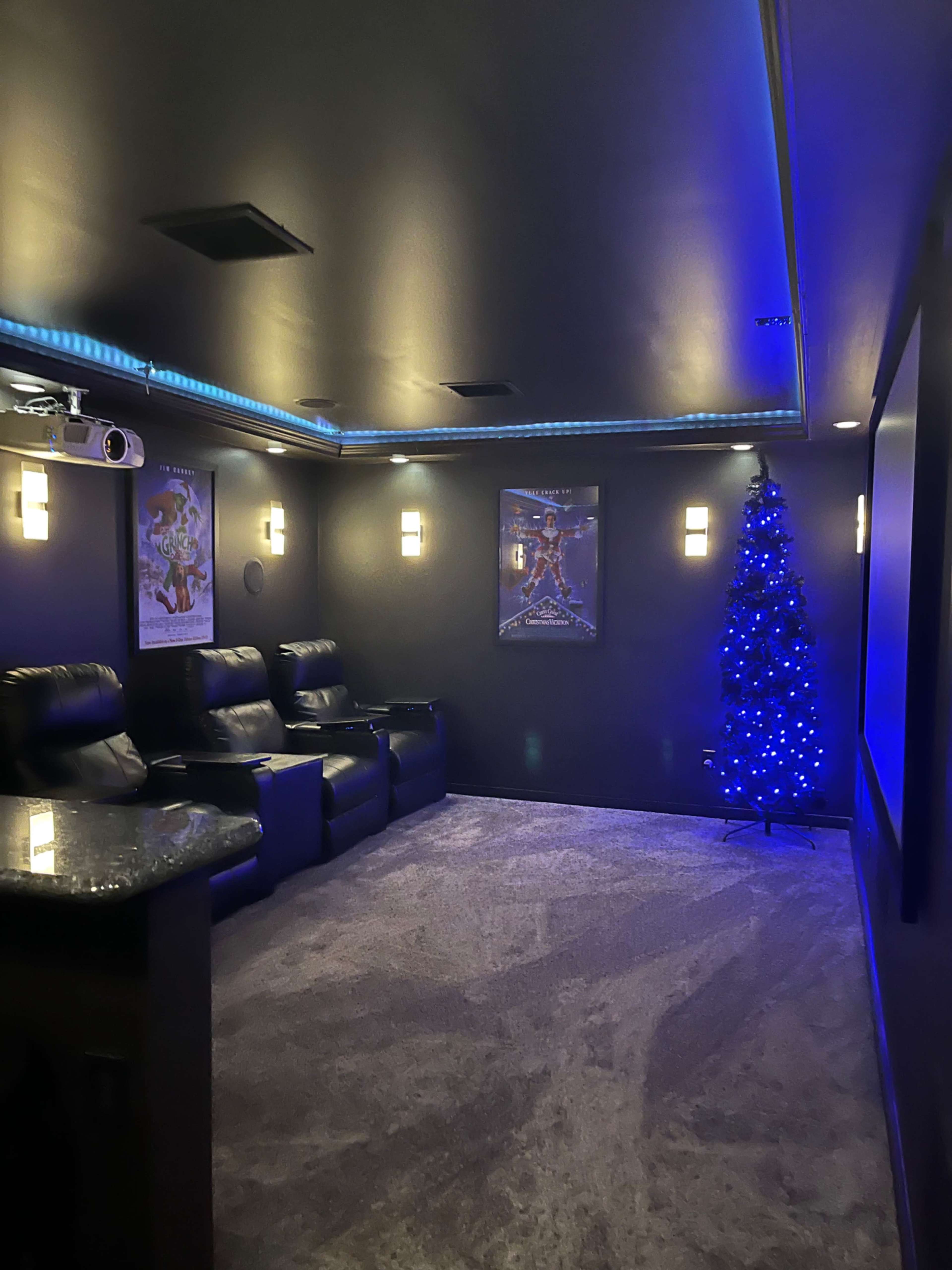 A home theater room with black recliner chairs, a Christmas tree with blue lights, and illuminated wall sconces.